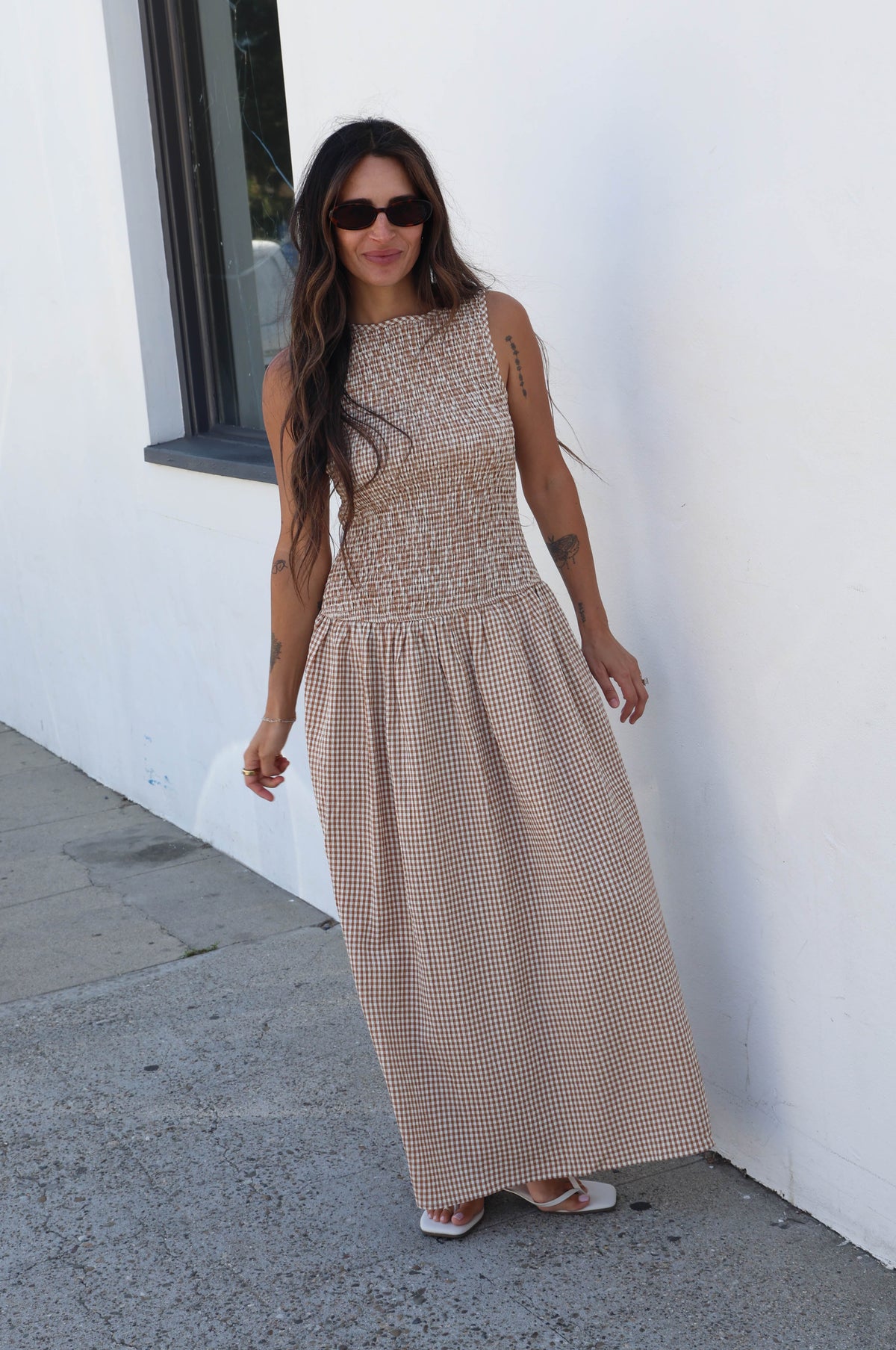 A woman wearing a sleeveless beige maxi dress with a textured bodice and a subtle patterned skirt, standing outdoors against a white wall.