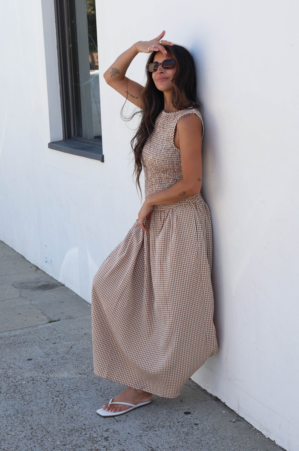 A woman wearing a sleeveless beige and white gingham maxi dress leans against a white stucco wall on a sunny day, wearing sunglasses and white flip-flops.