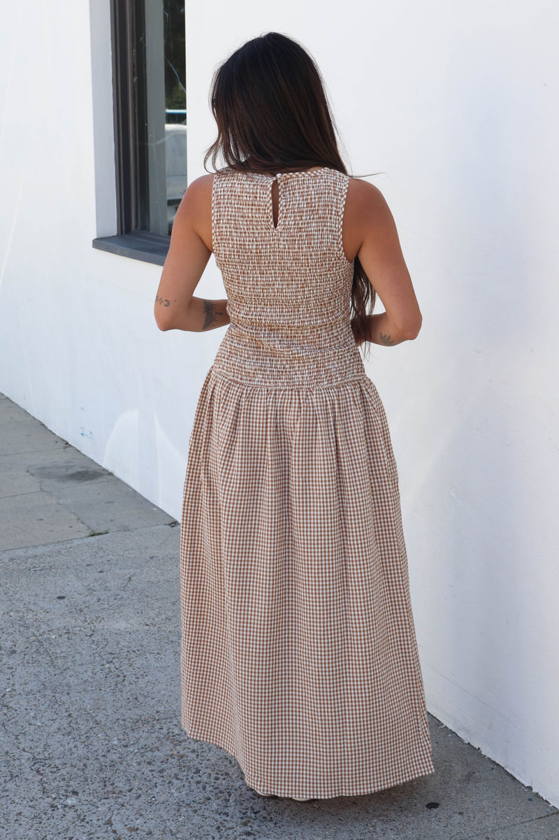 A woman viewed from behind wearing a sleeveless, smocked maxi dress with a beige and white gingham pattern, standing against a white stucco wall.