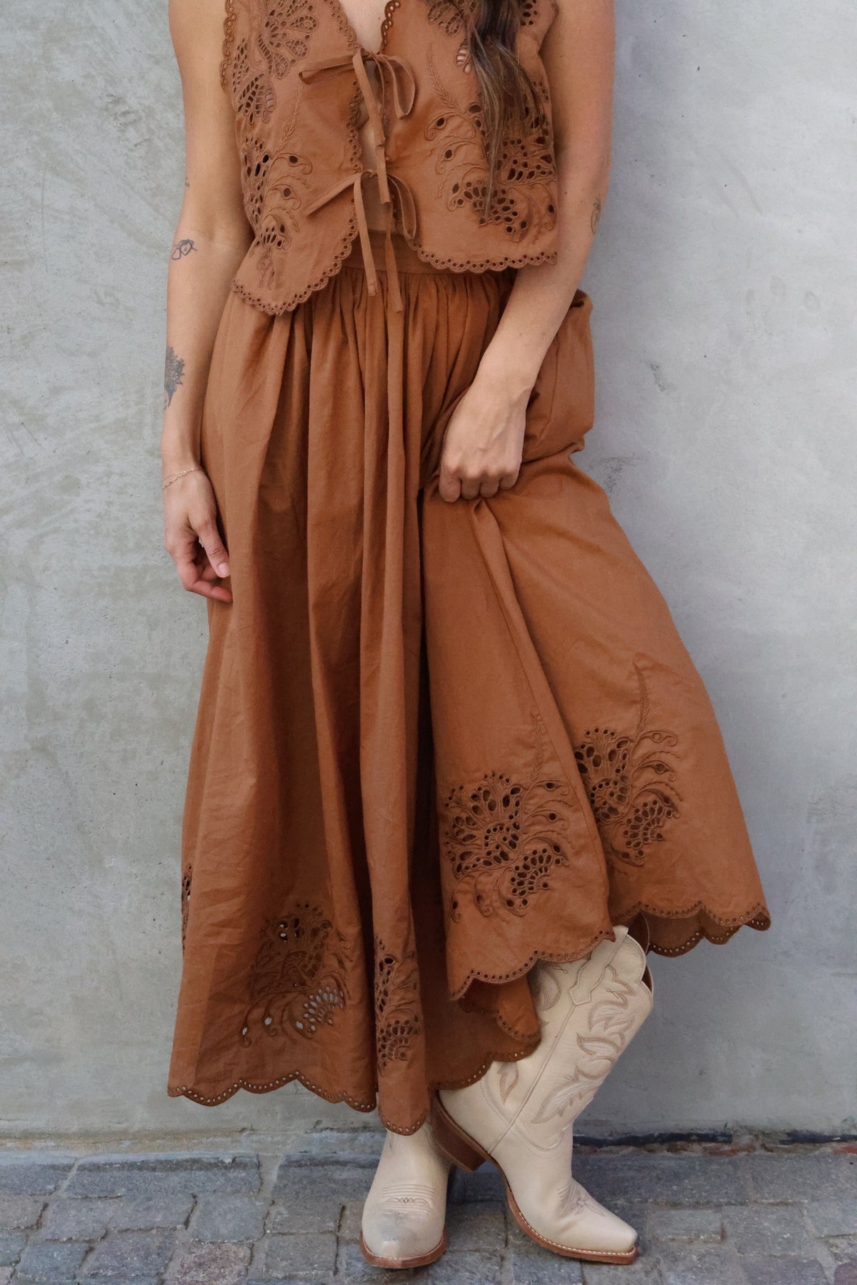 Person wearing a long brown dress with lace details against a gray wall.