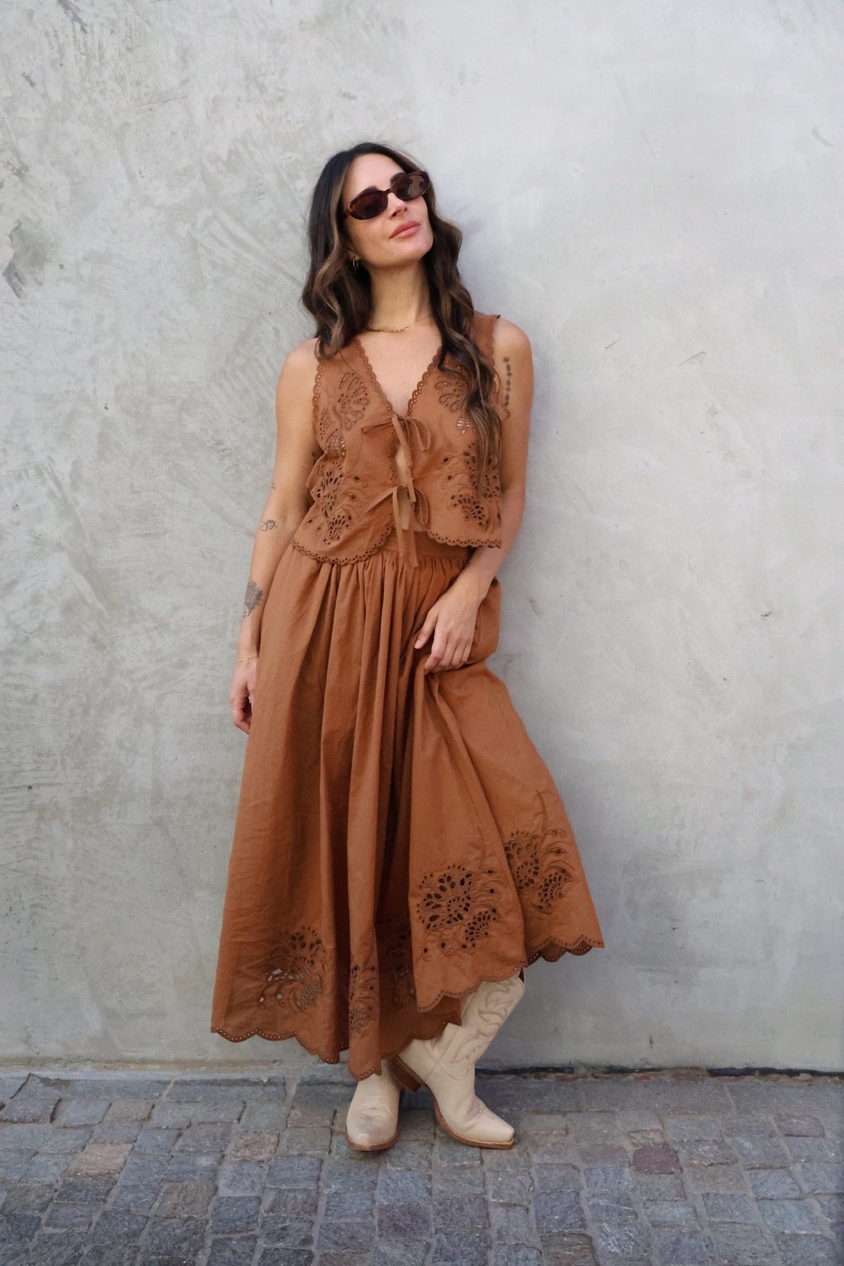Woman wearing a brown lace dress and vest against a gray wall.