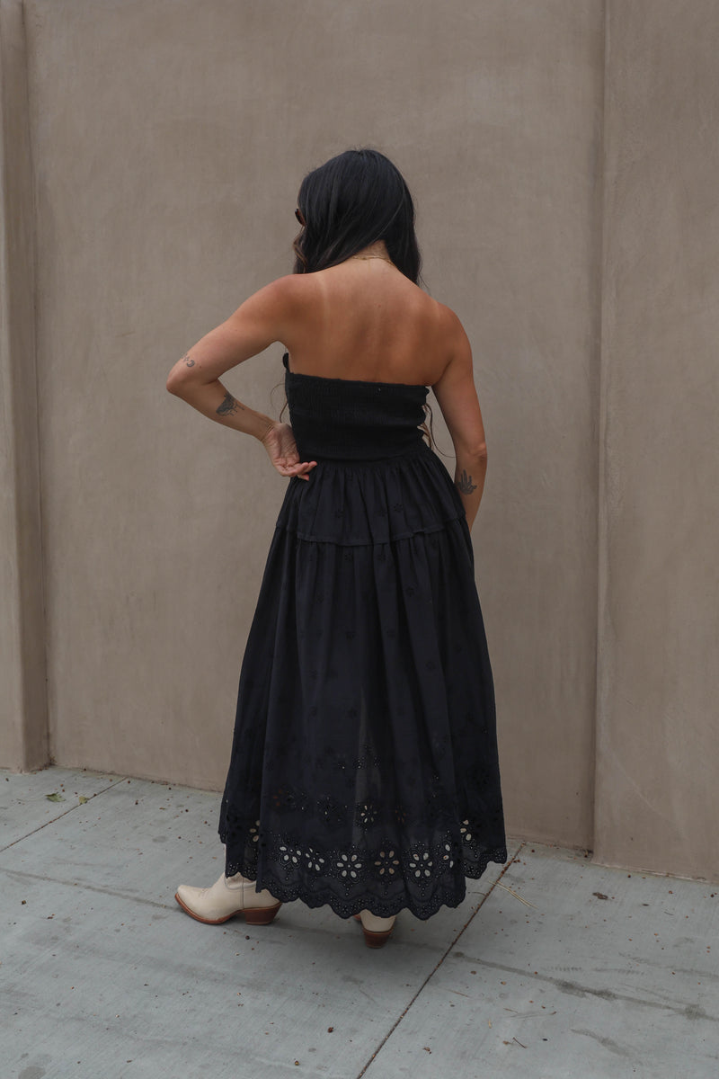 Person wearing a black strapless dress with floral details, standing against a beige wall.