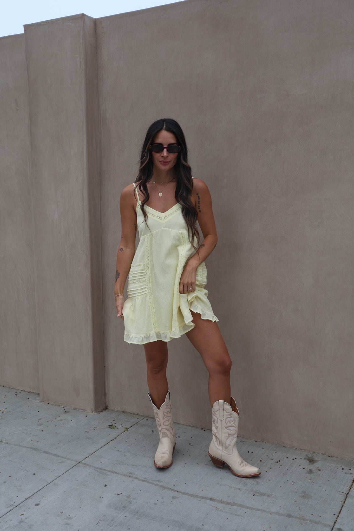 Woman in a light yellow dress and white cowboy boots standing against a plain wall.