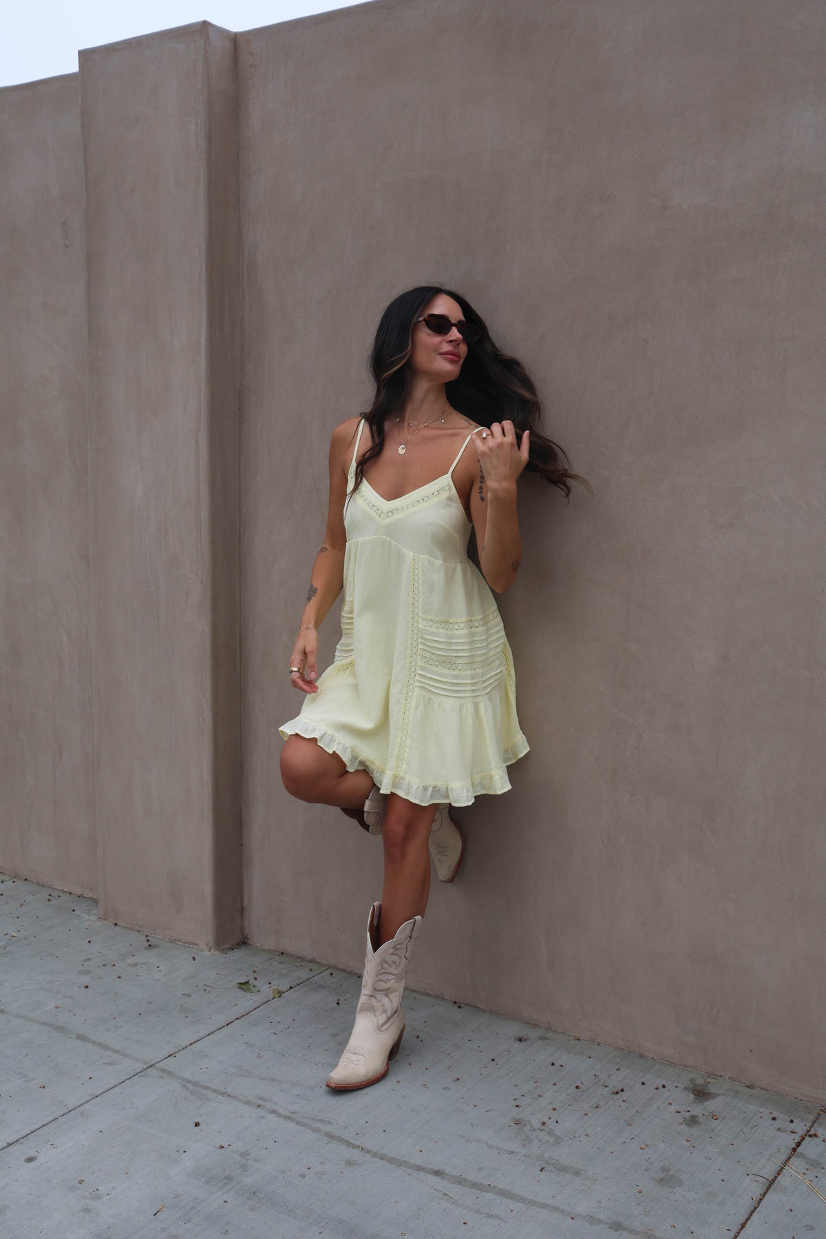 Woman in a light yellow dress and white boots leaning against a beige wall.