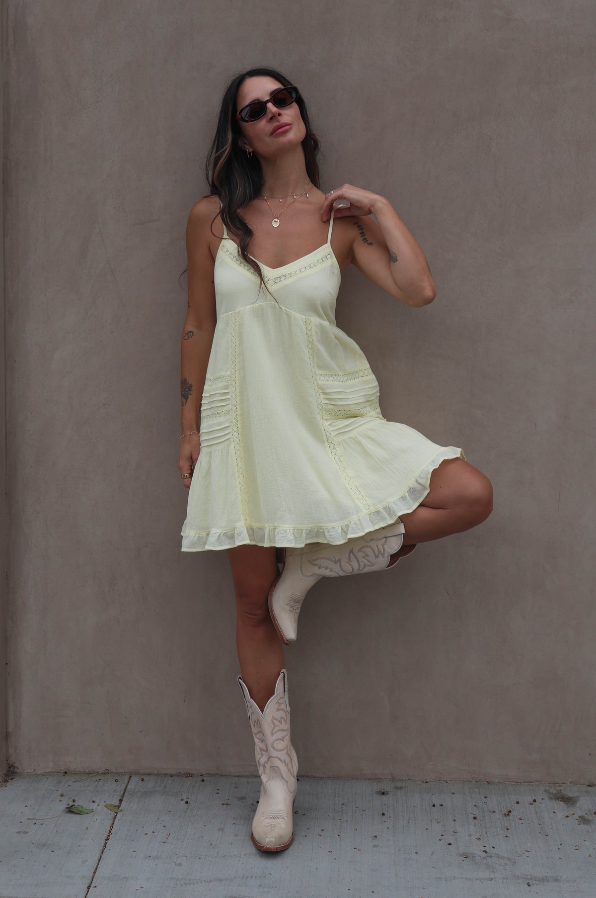 Woman in a light yellow dress and white boots posing against a plain wall.