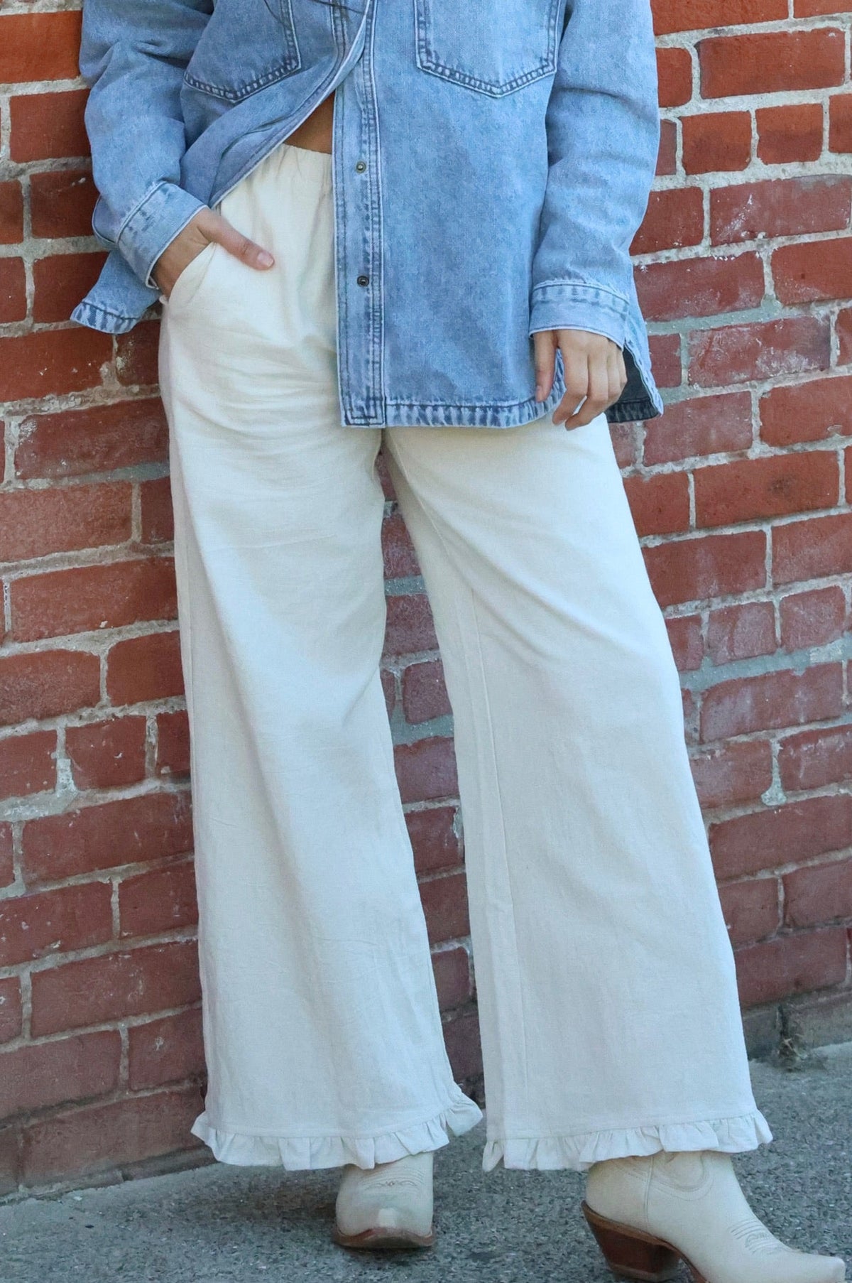 Person wearing a denim jacket and white pants standing against a brick wall.