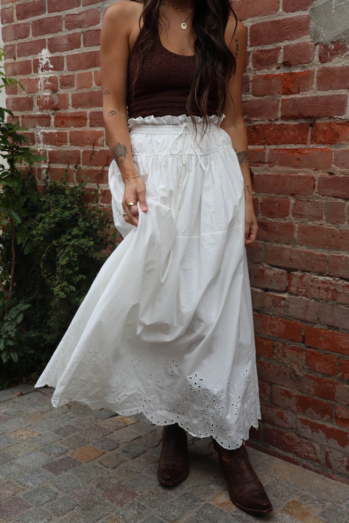 Person wearing a white lace skirt and brown top against a brick wall.