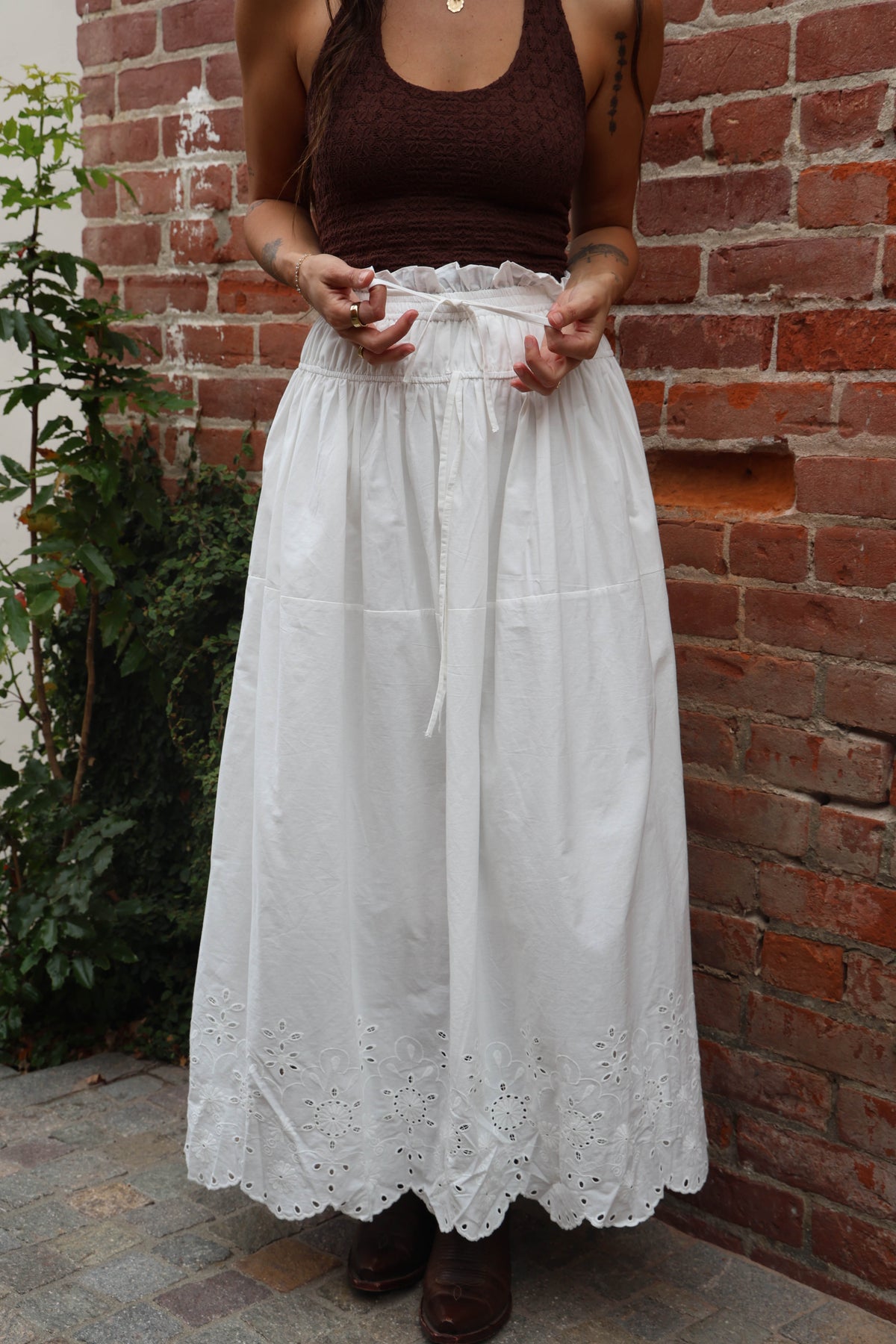 Person wearing a brown tank top and white lace skirt against a brick wall.