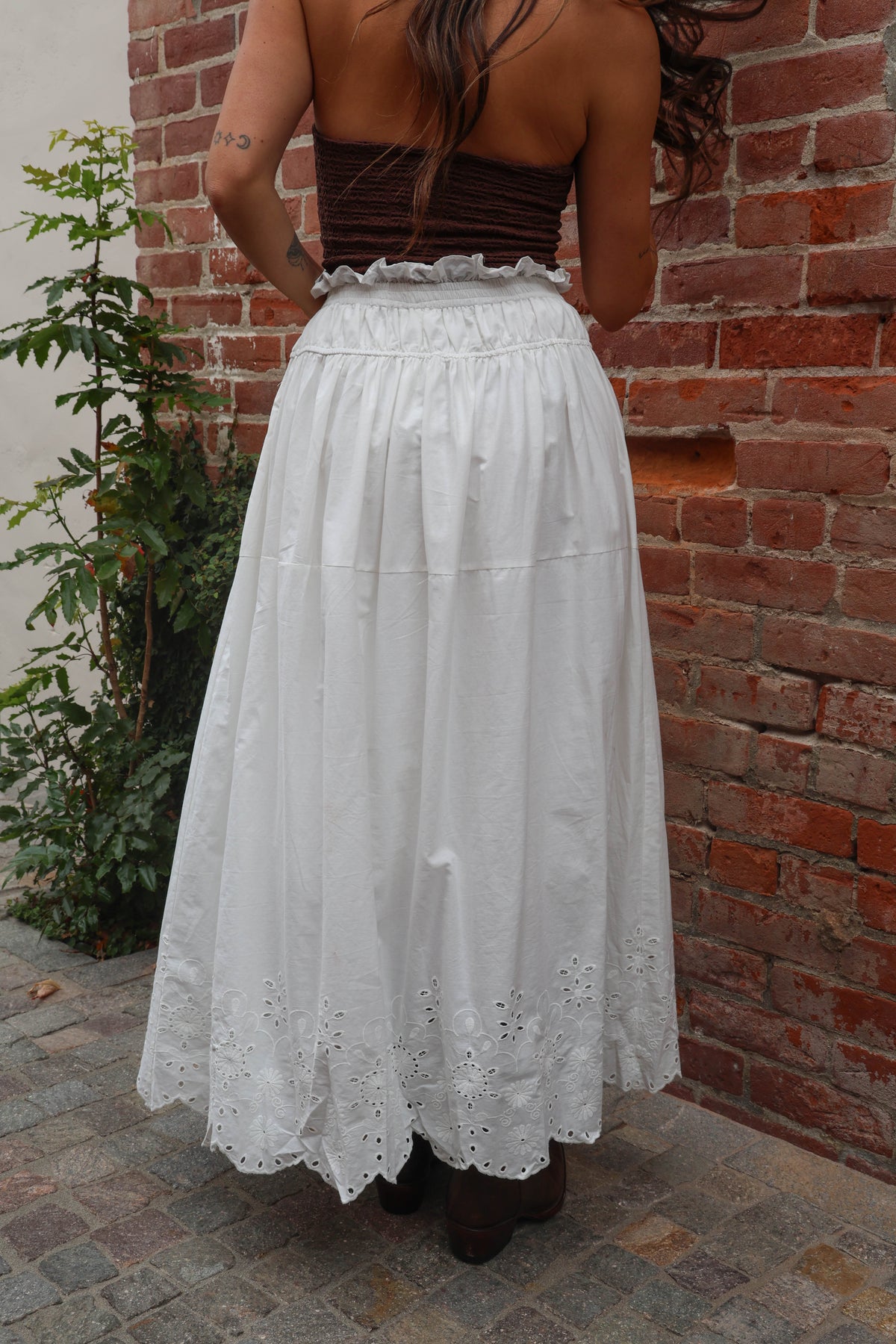 Person wearing a white lace skirt and brown top against a brick wall.