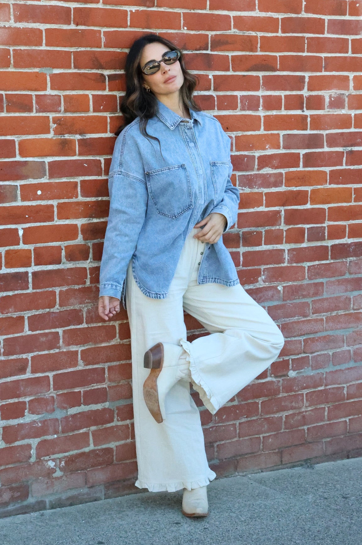 Woman in denim shirt and white pants leaning against a brick wall.