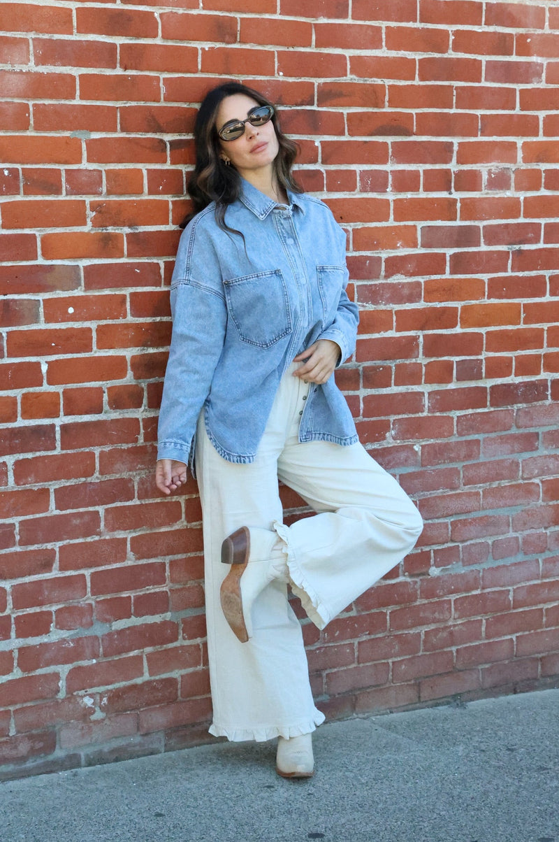 Woman in denim shirt and white pants leaning against a brick wall.