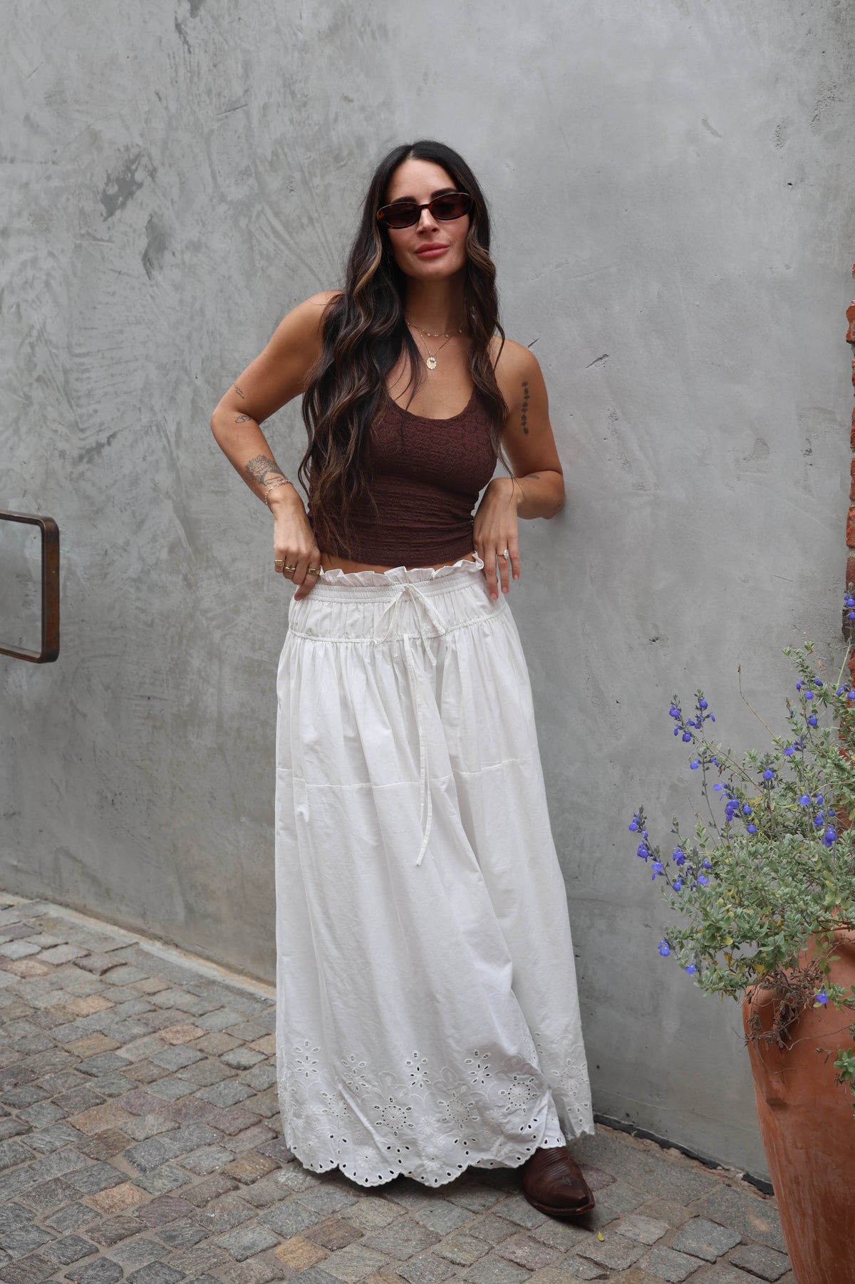 Woman wearing a brown top and white lace skirt standing against a gray wall.