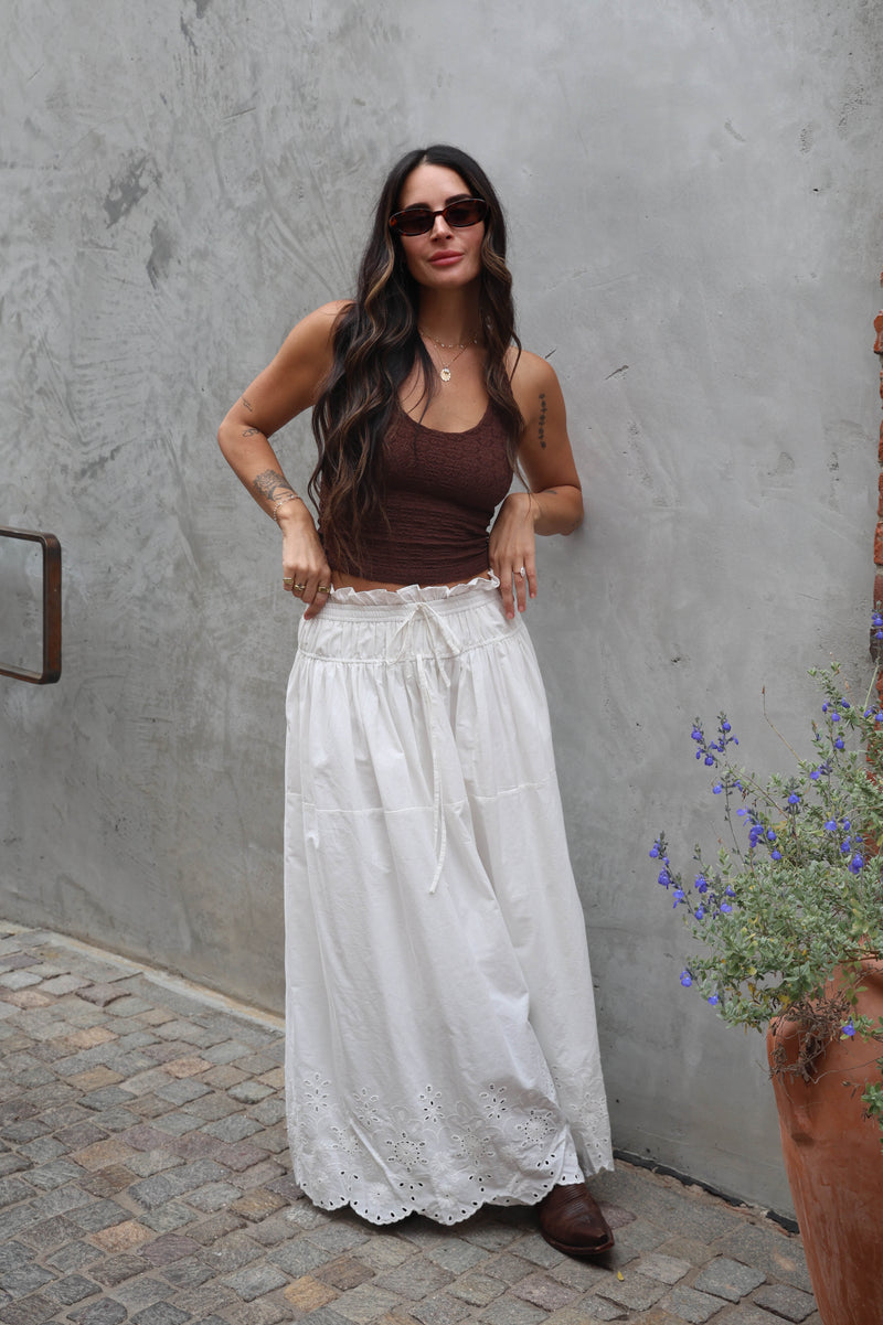 Woman wearing a brown top and white lace skirt standing against a gray wall.