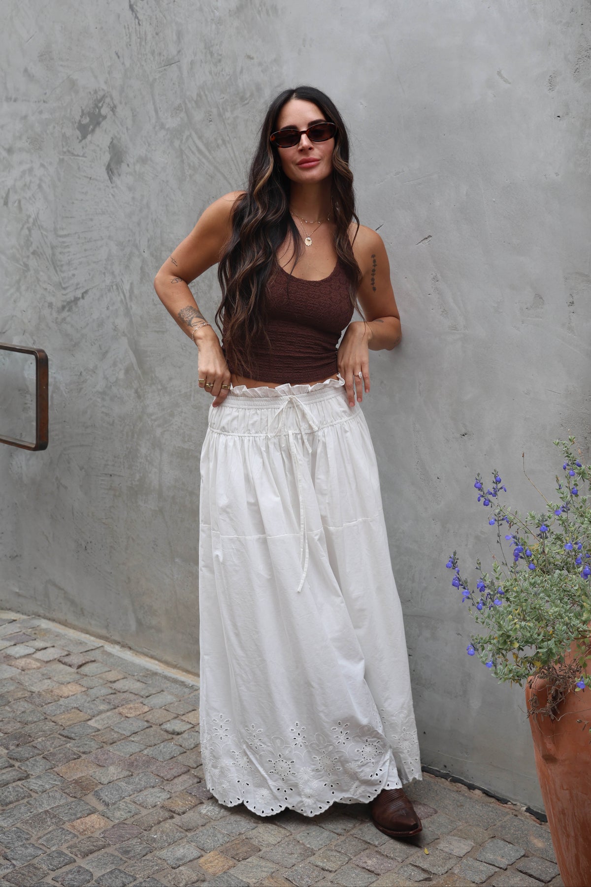 Woman wearing a brown top and white lace skirt standing against a gray wall.