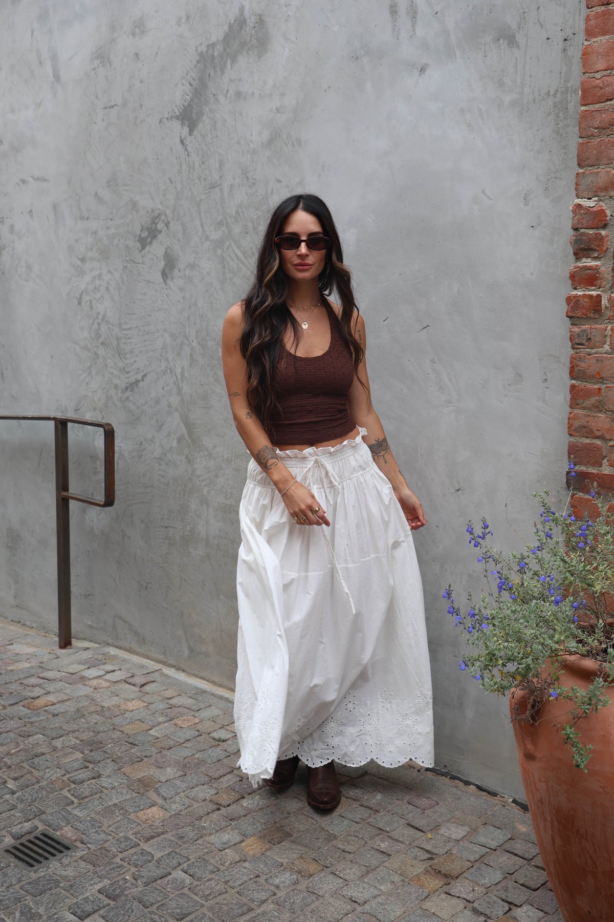 Woman in a brown top and white skirt standing against a gray wall with a brick column.