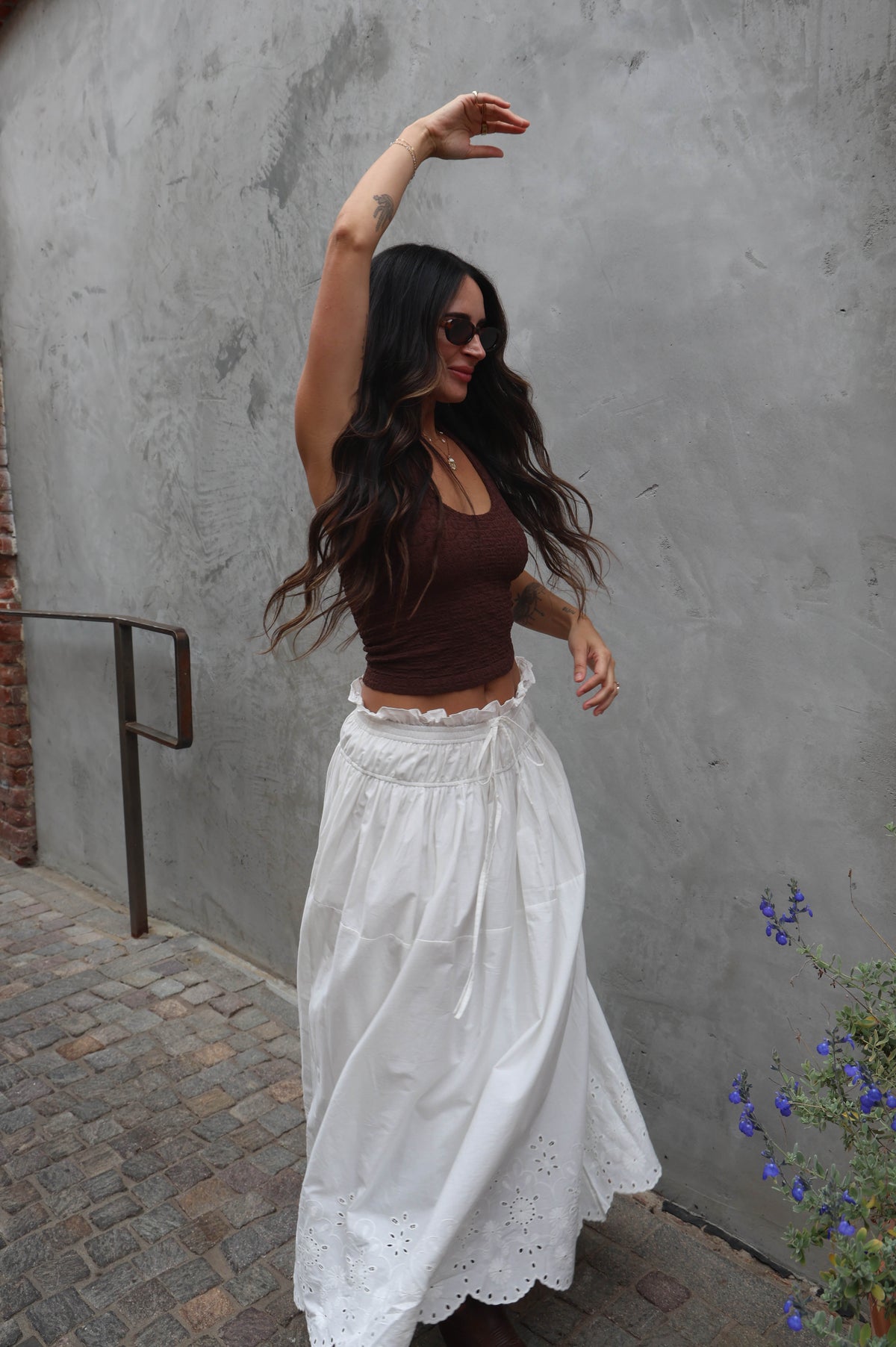 Woman in a brown top and white skirt standing against a gray wall.