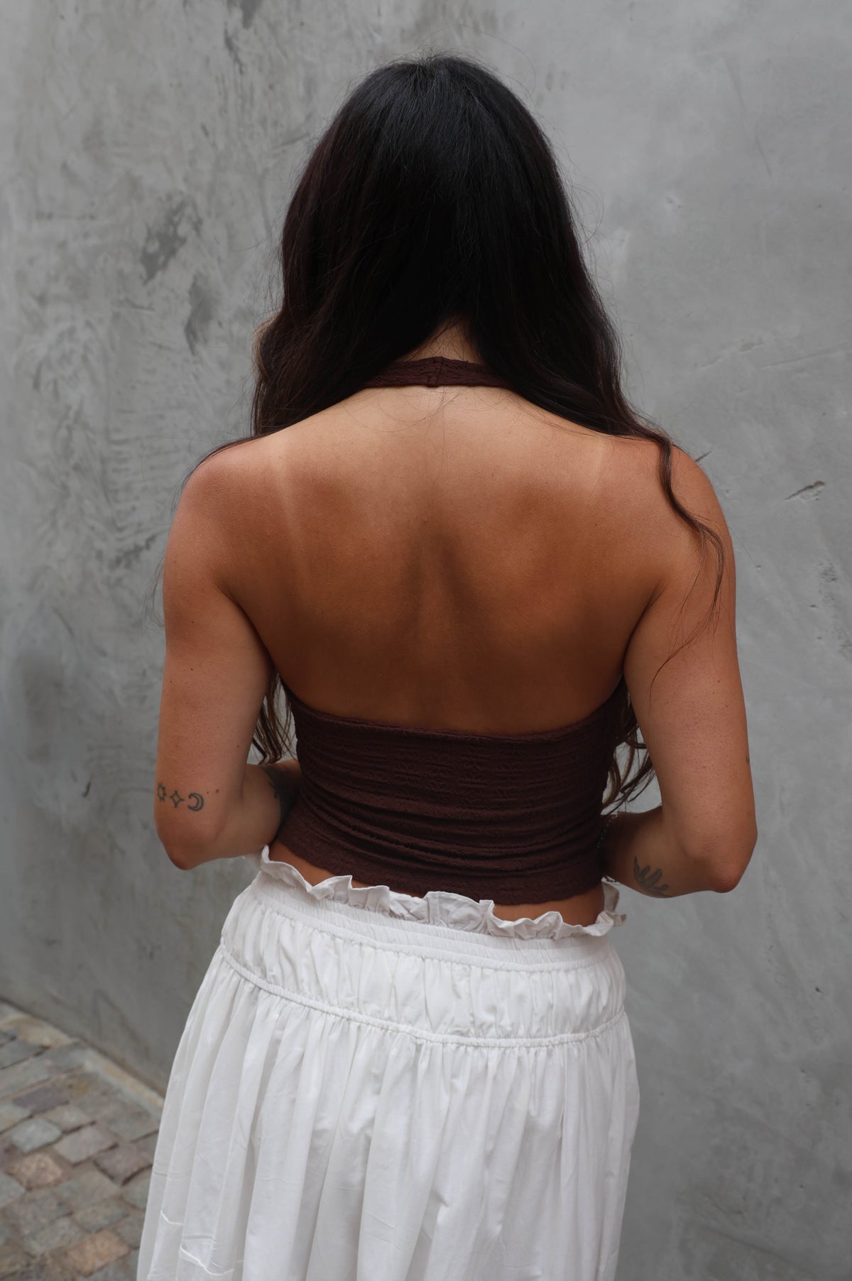 Woman wearing a brown top and white pants against a gray wall.