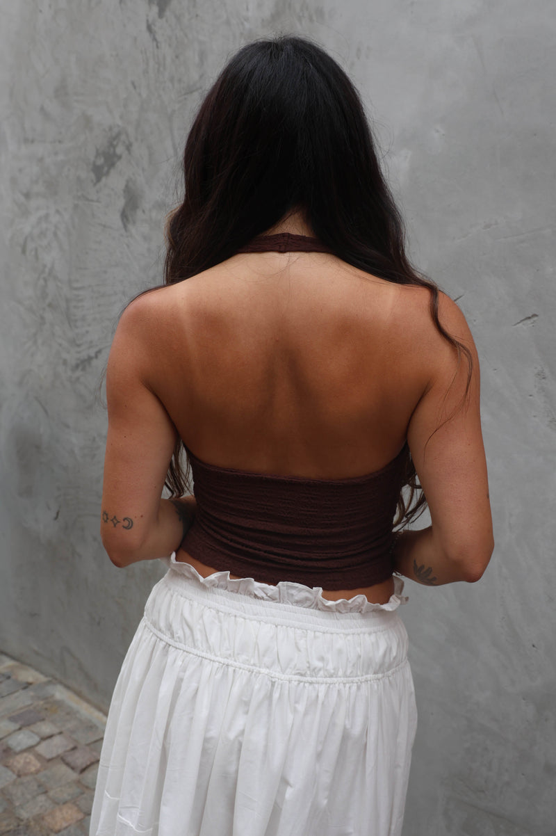 Woman wearing a brown top and white pants against a gray wall.
