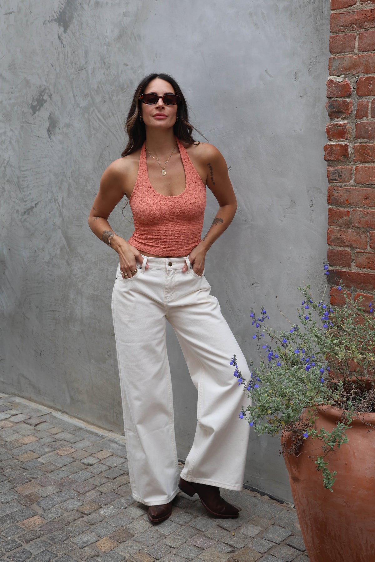 Woman in a coral top and white pants standing against a gray wall with a potted plant.