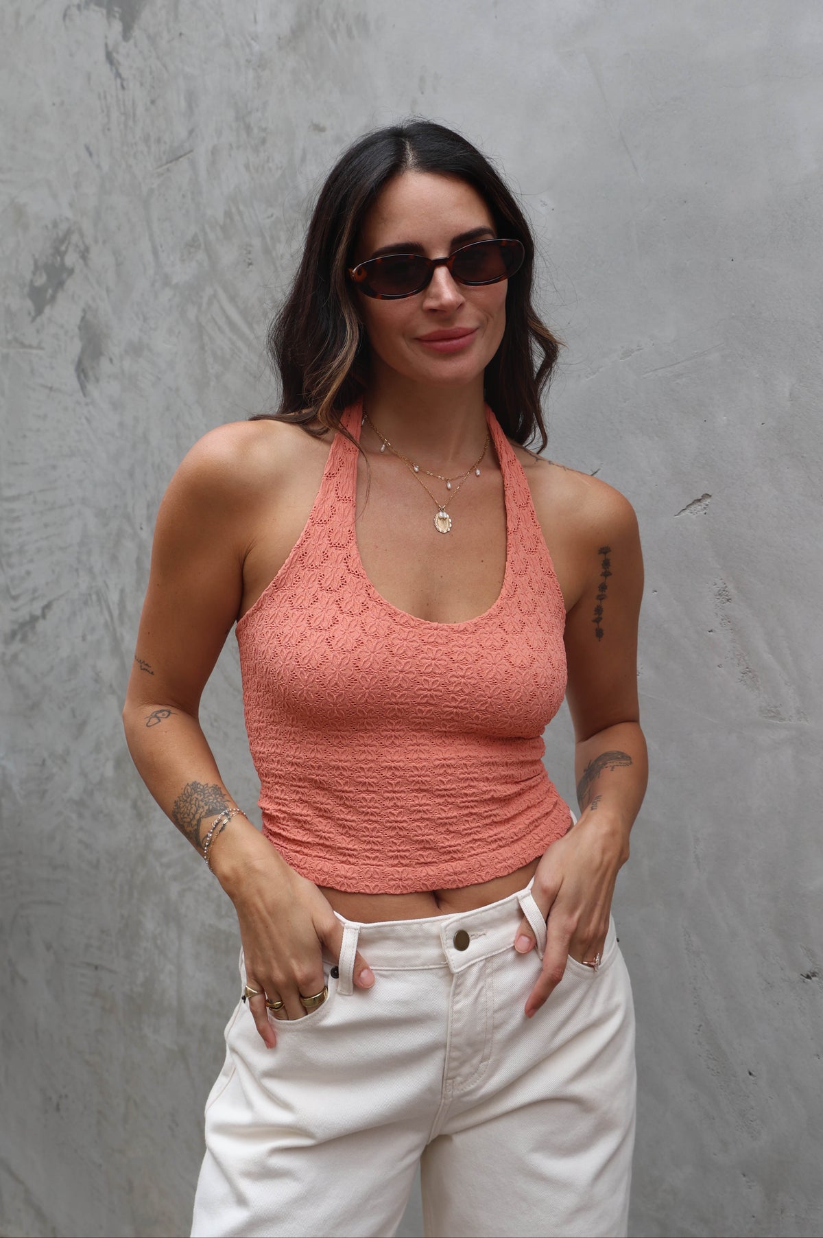Woman wearing a coral tank top and white pants against a gray wall