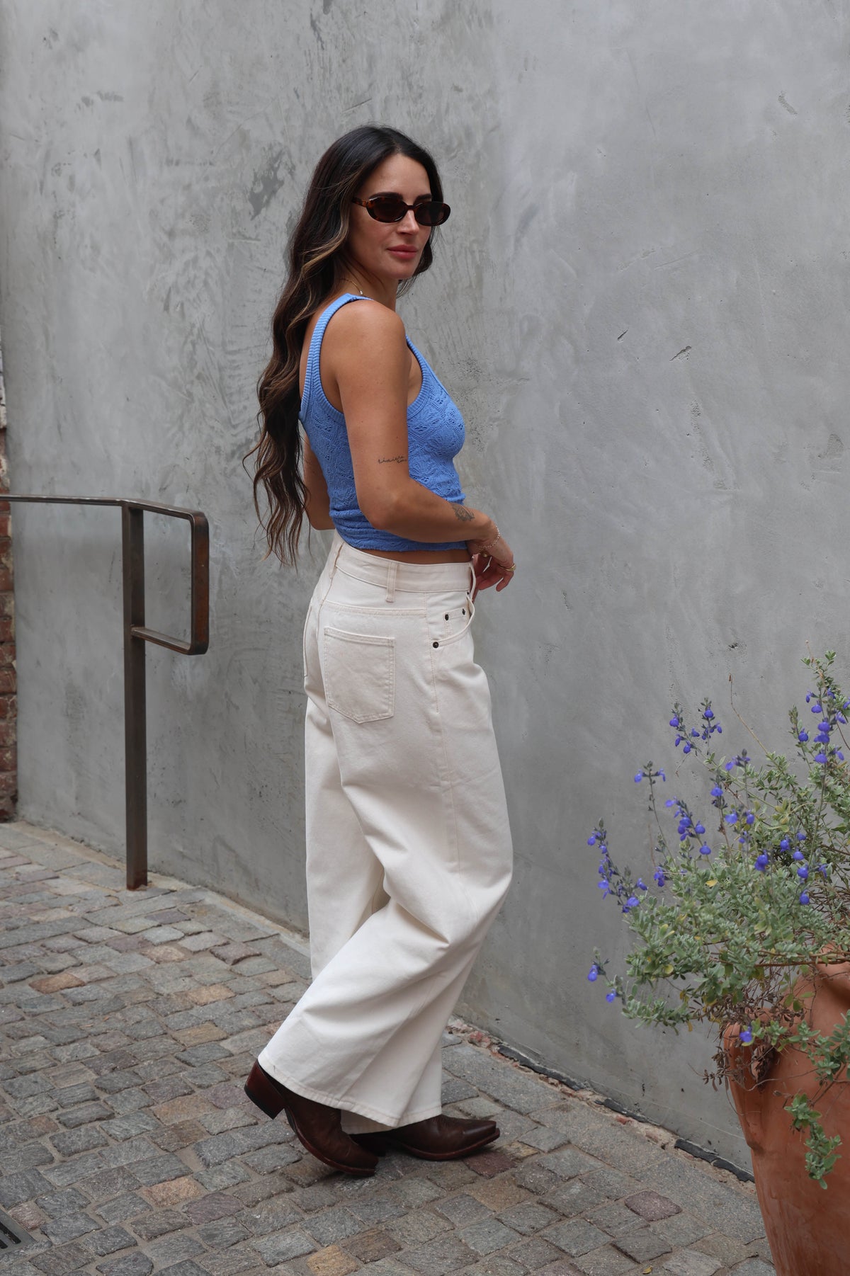 Woman in blue top and white pants standing against a gray wall with a potted plant nearby.