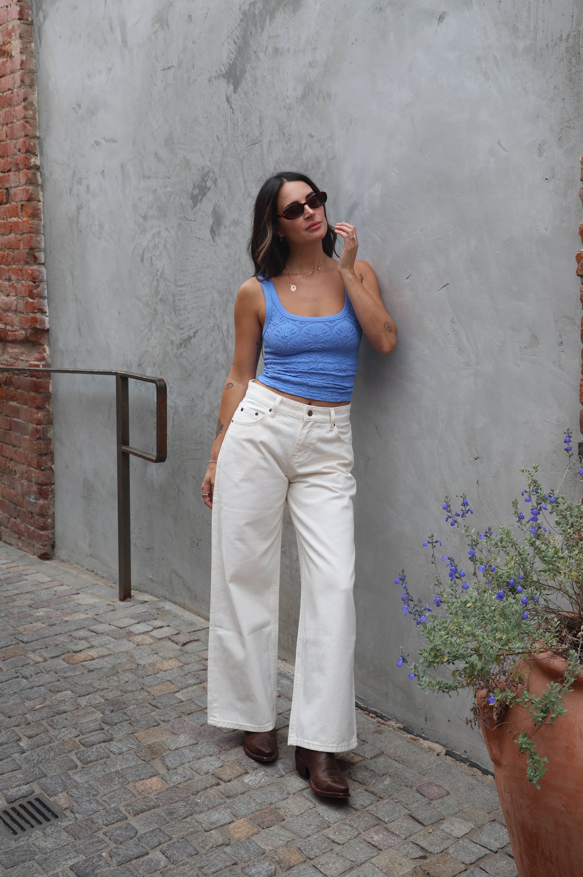 Woman in blue top and white pants standing against a gray wall with a brick border.