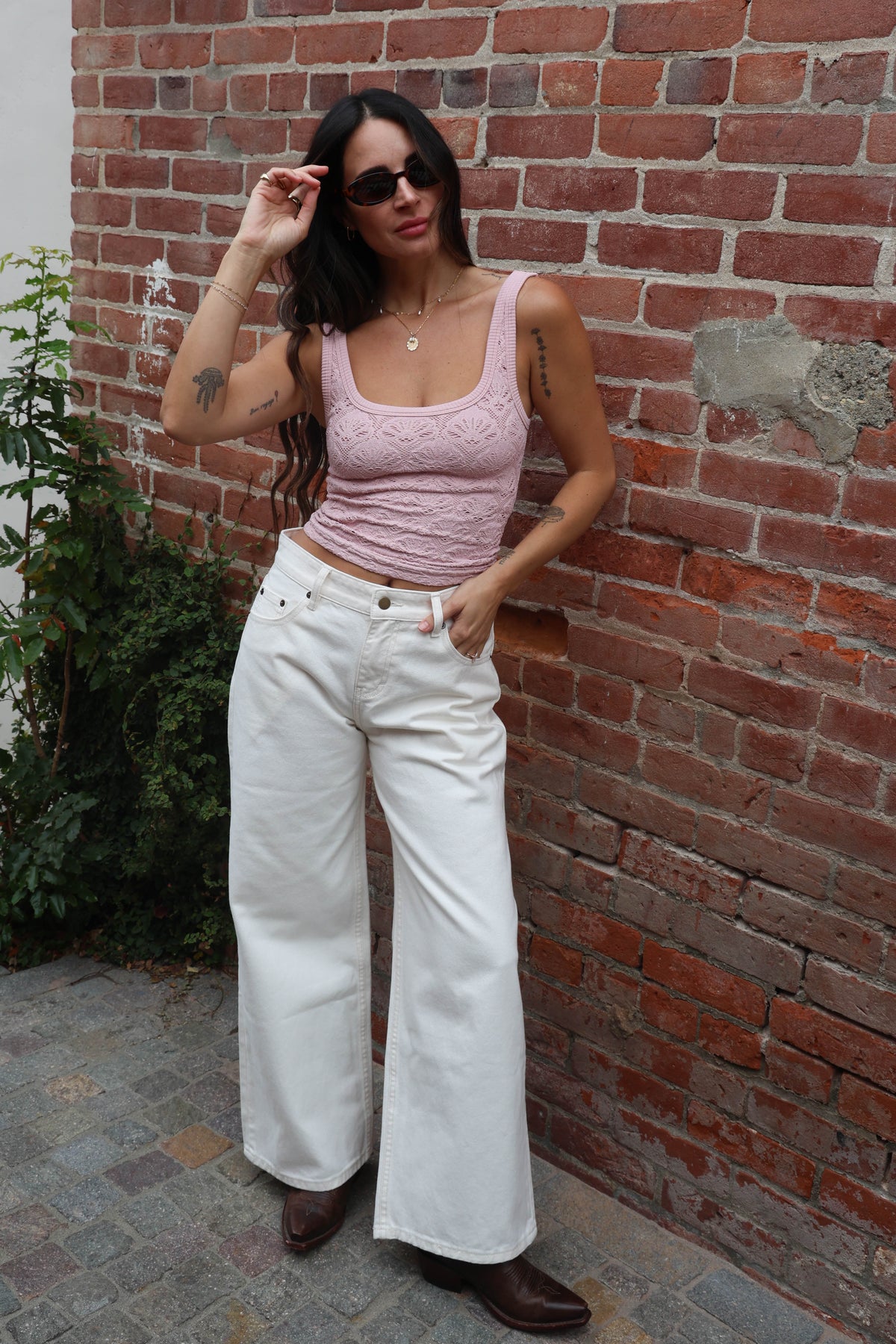 Woman in pink top and white pants standing against a brick wall.