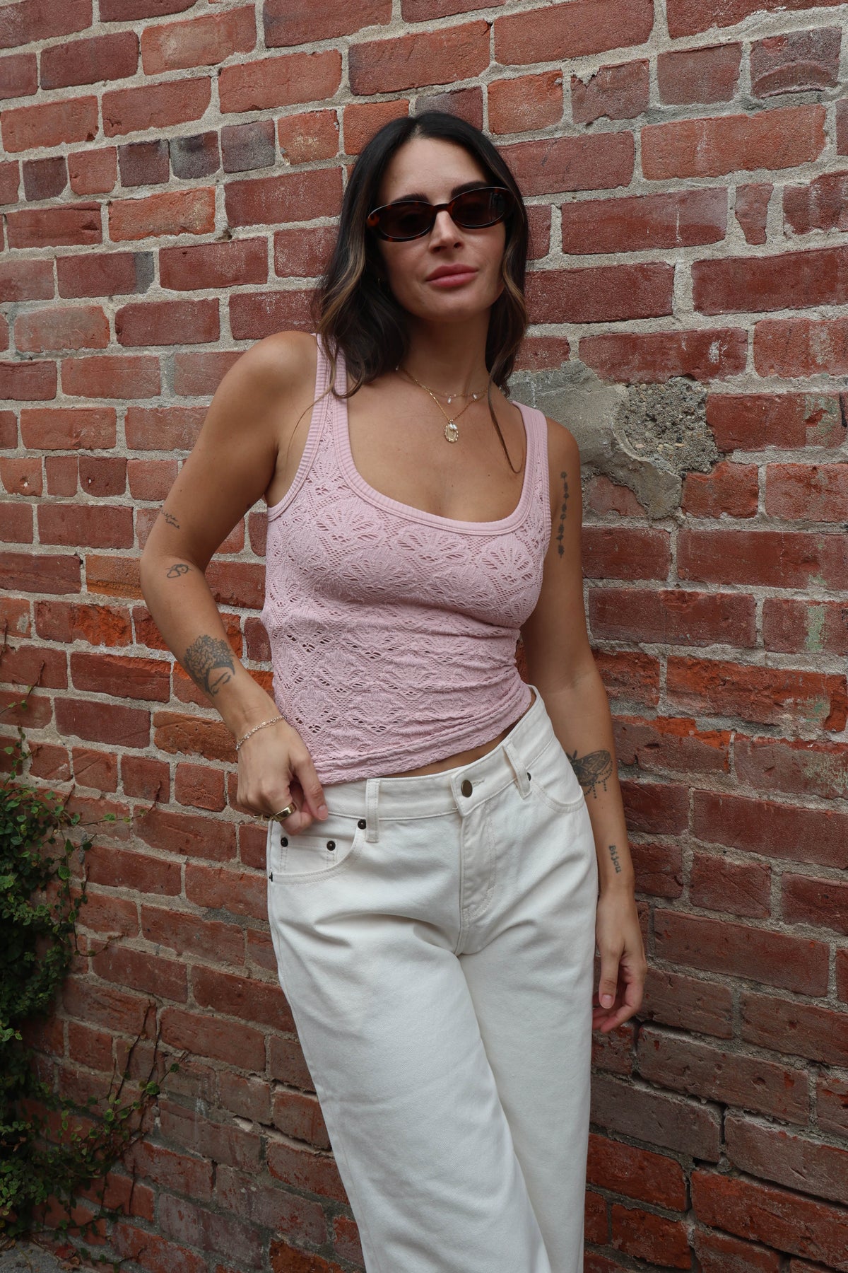 Woman wearing a pink tank top and white pants standing against a brick wall.