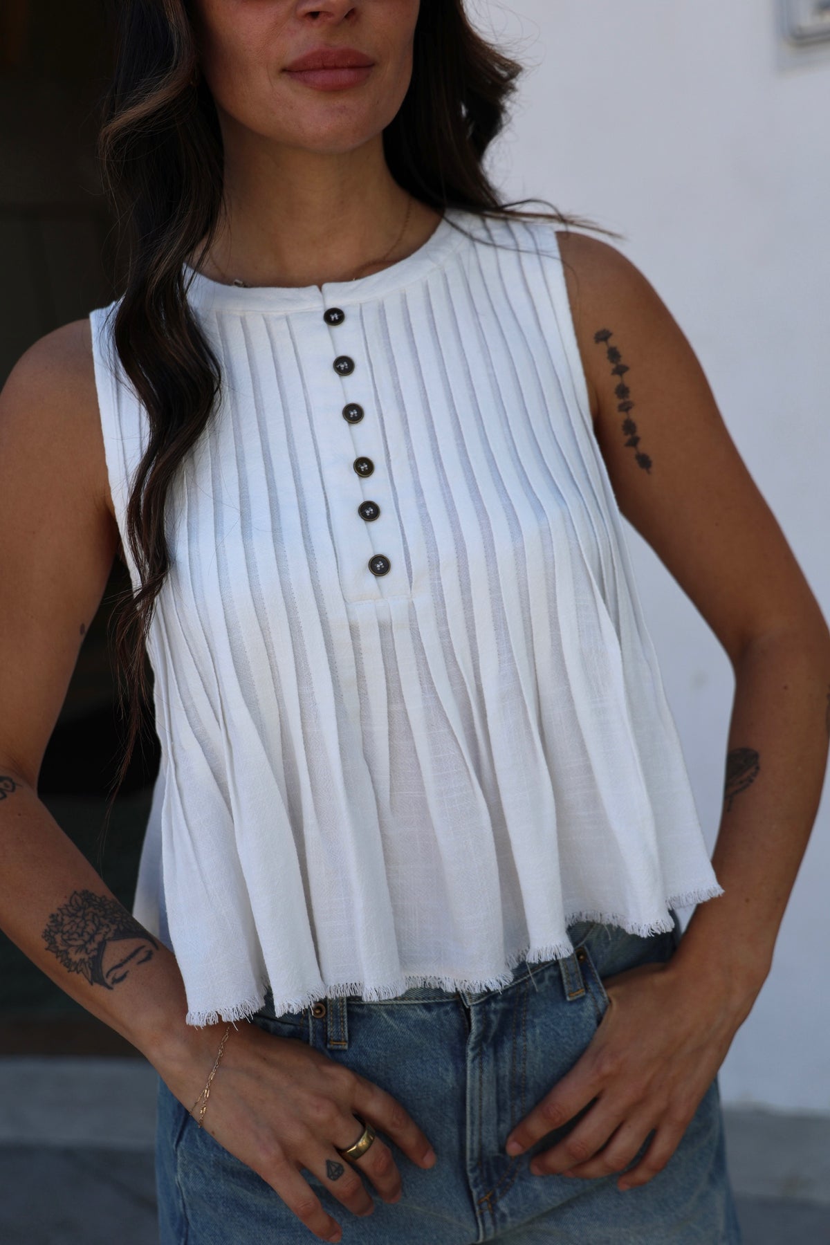 Woman wearing a white sleeveless top with black buttons and blue jeans.