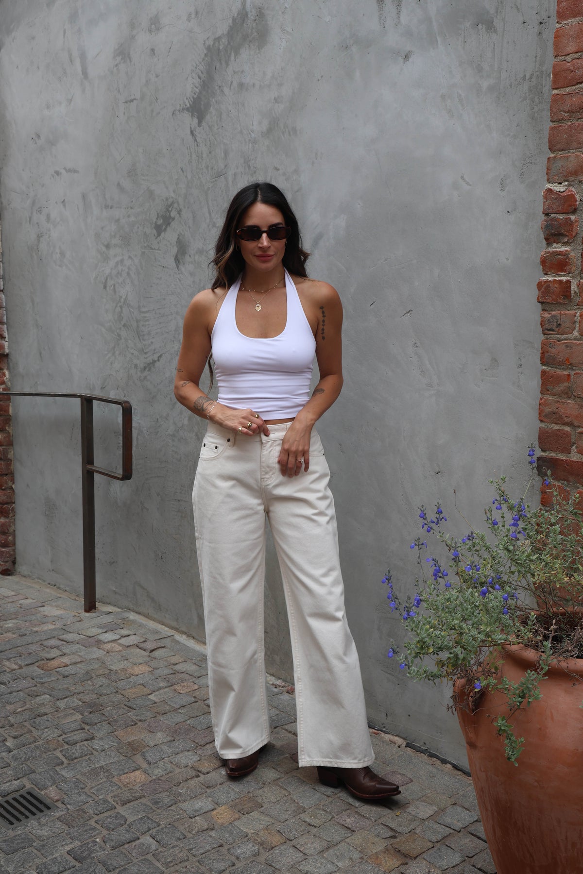 Woman in white tank top and pants standing against a gray wall with a plant nearby.