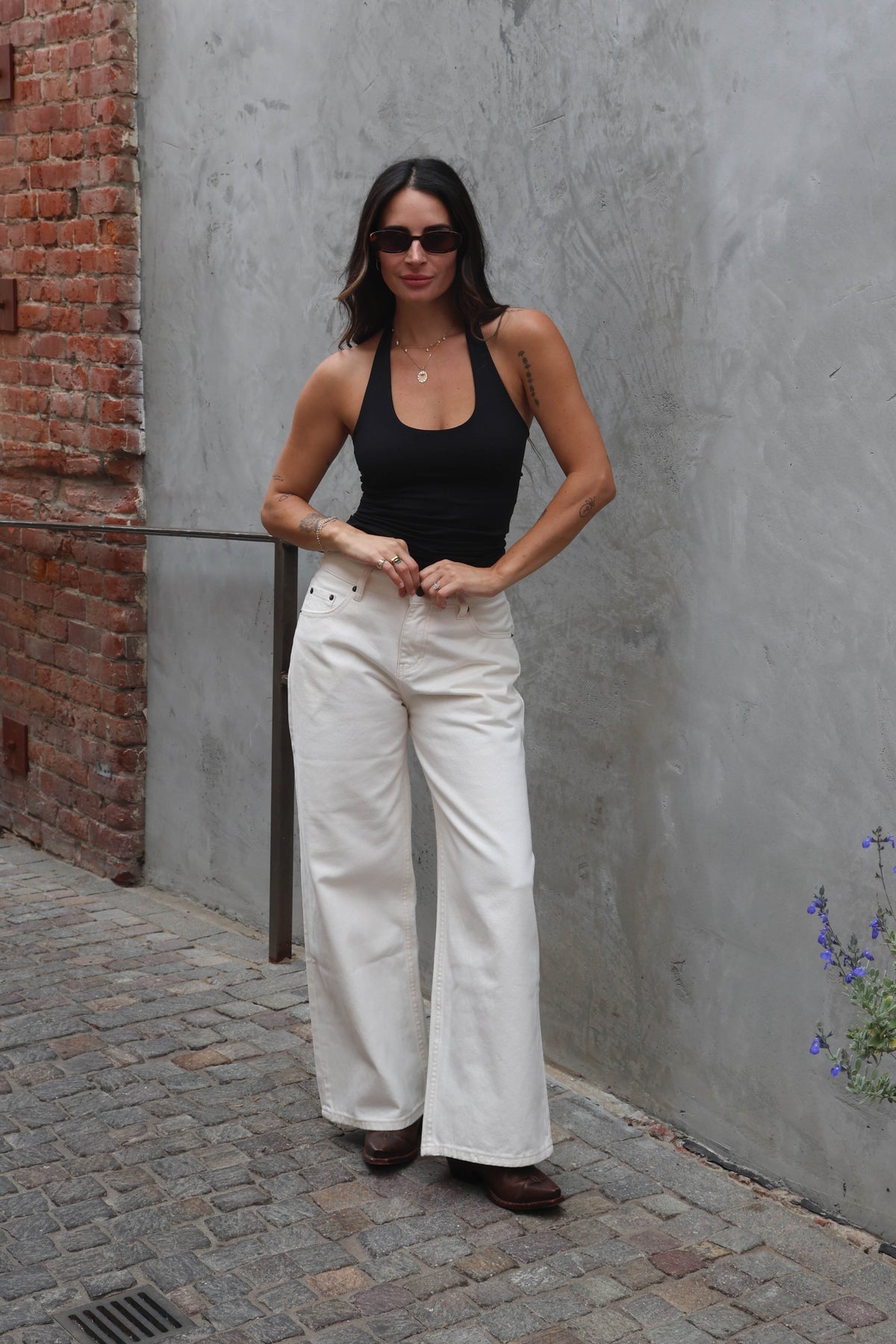 Woman wearing a black tank top and white pants standing against a gray wall.
