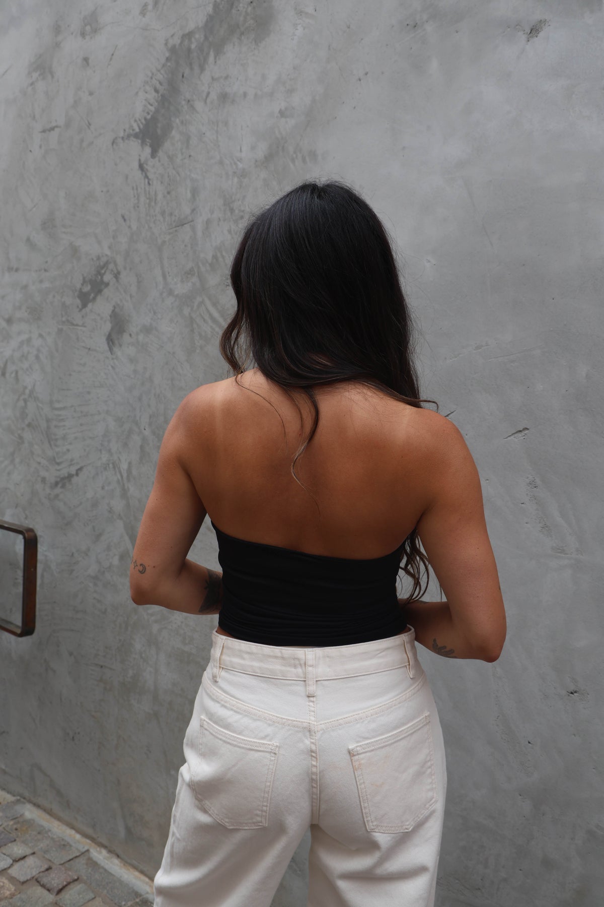 Person wearing a black top and white pants against a gray wall