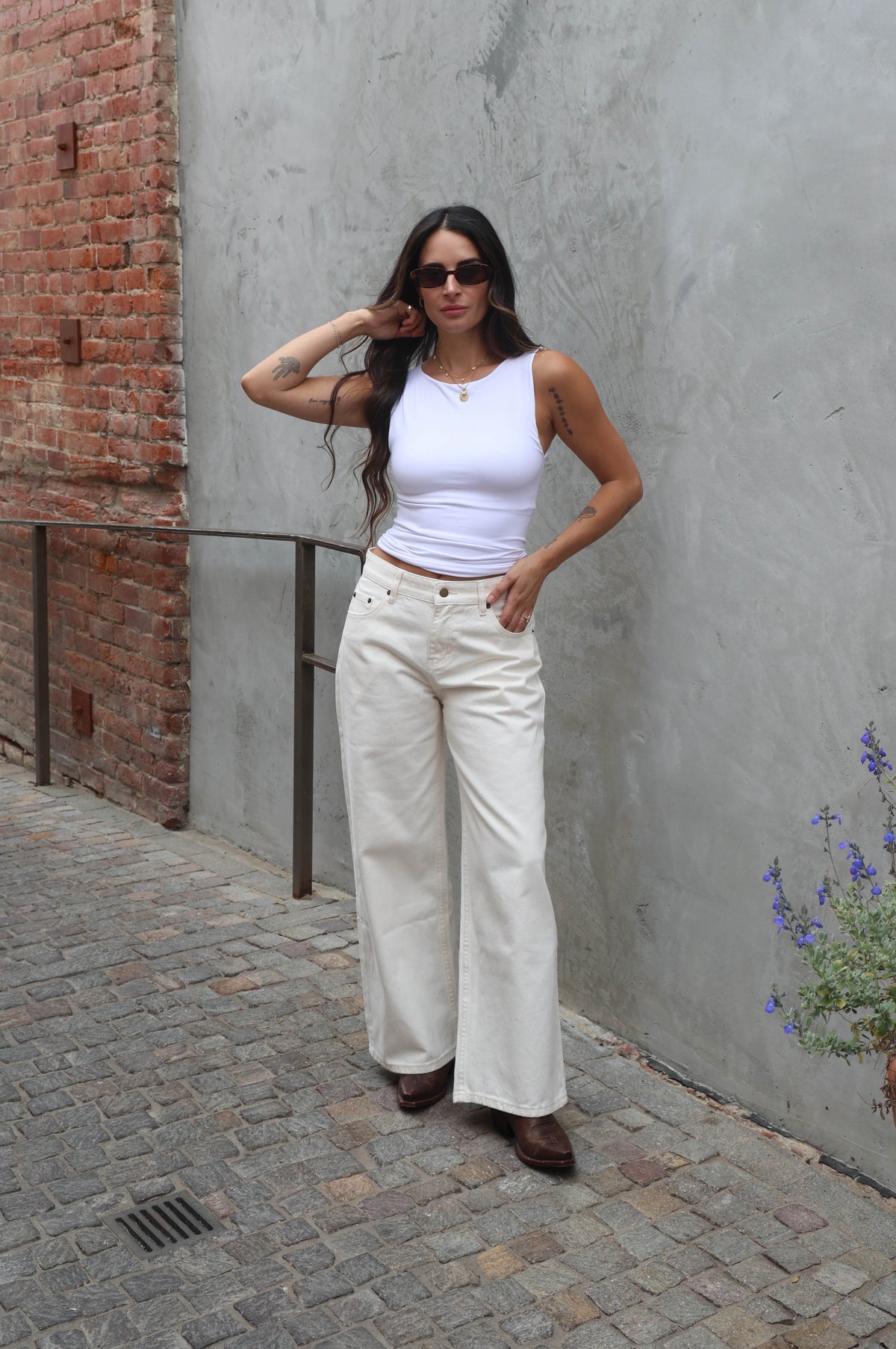 Woman in white top and pants standing against a gray wall with a brick wall in the background.