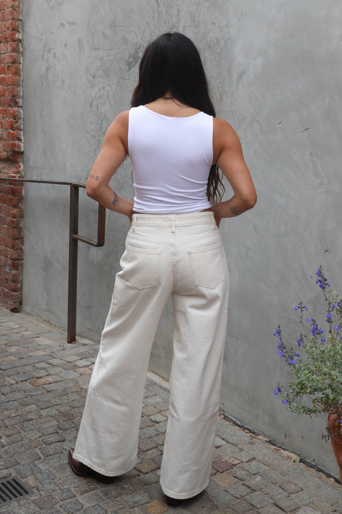 Person wearing a white sleeveless top and cream-colored pants standing against a gray wall.