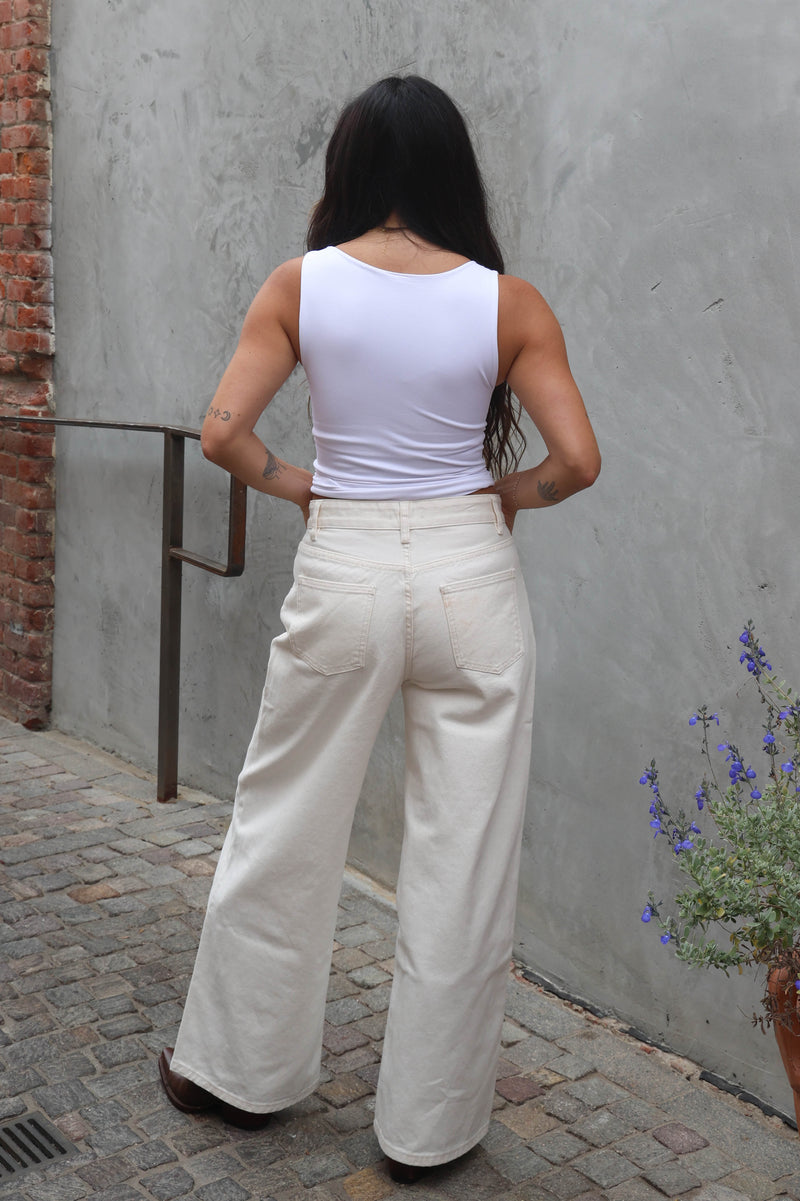 Person wearing a white sleeveless top and cream-colored pants standing against a gray wall.