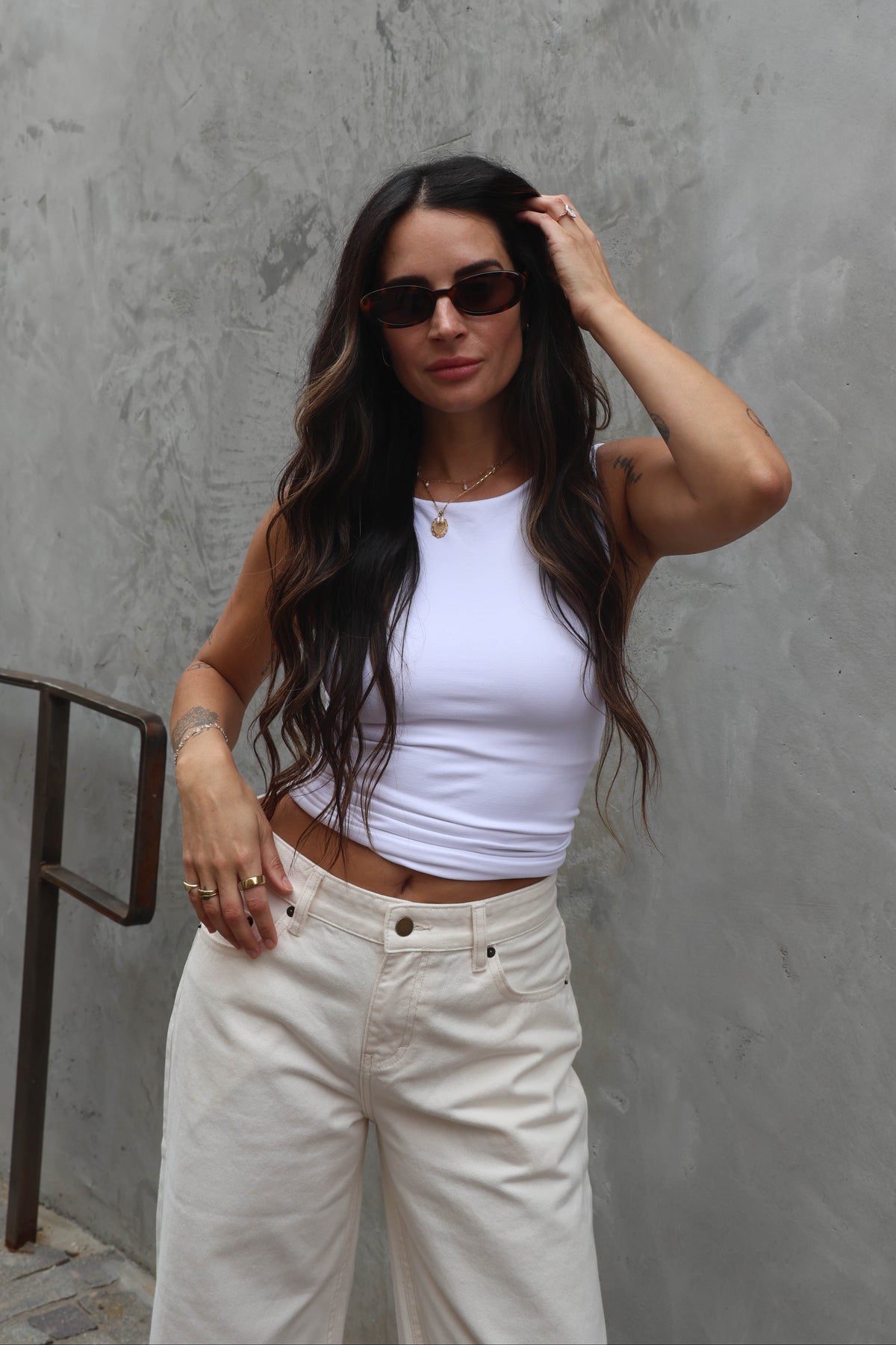 Woman wearing a white tank top and beige pants against a gray wall.