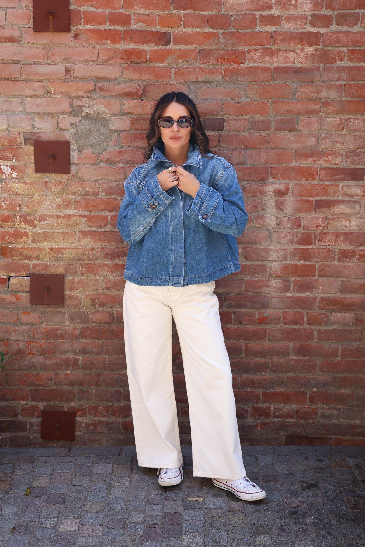A woman stands in front of a red brick wall wearing a light blue cropped denim jacket, off-white wide-leg trousers, and white sneakers.