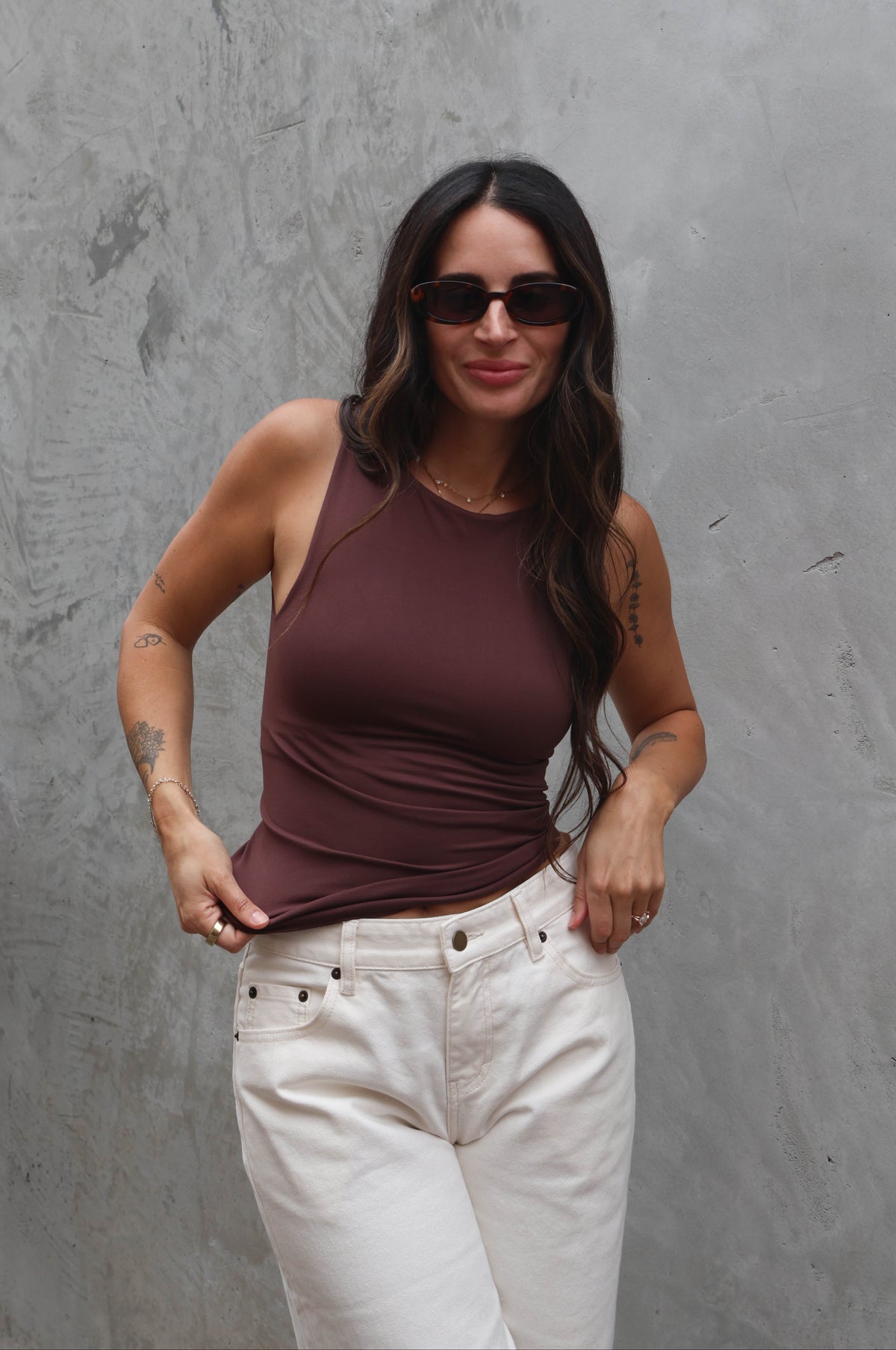 Woman wearing a brown tank top and white pants against a gray wall.
