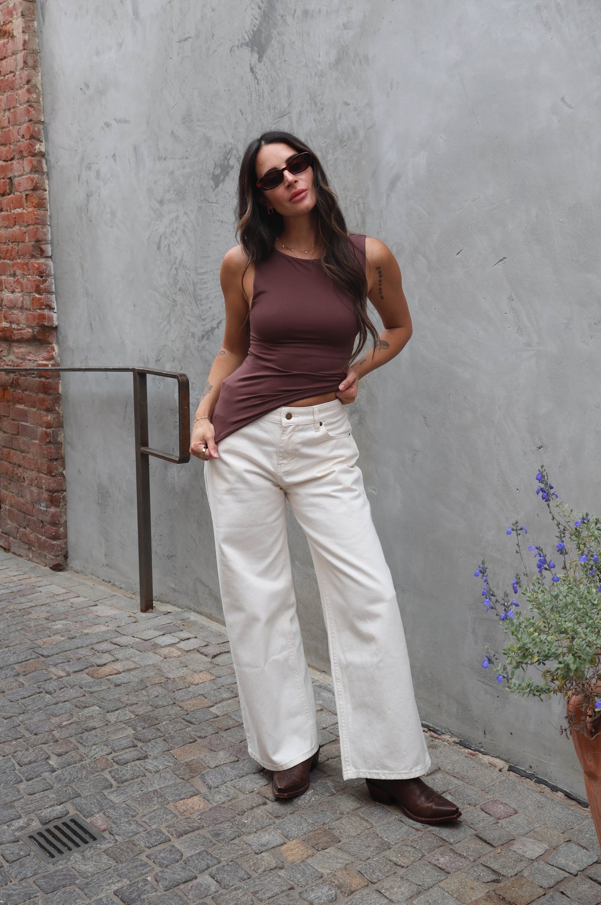 Woman in a brown tank top and white pants standing against a gray wall.