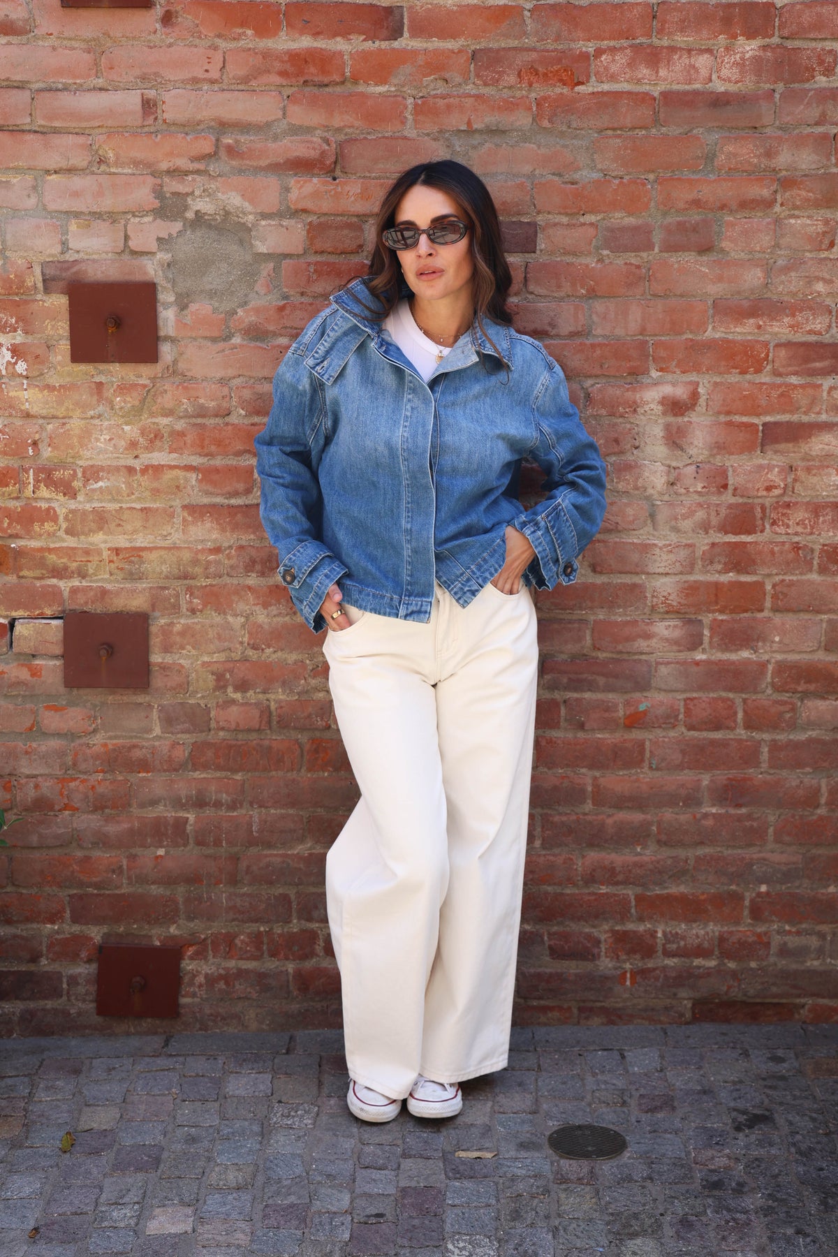 A woman wearing a blue denim jacket, white wide-leg trousers, and white sneakers stands against a red brick wall.