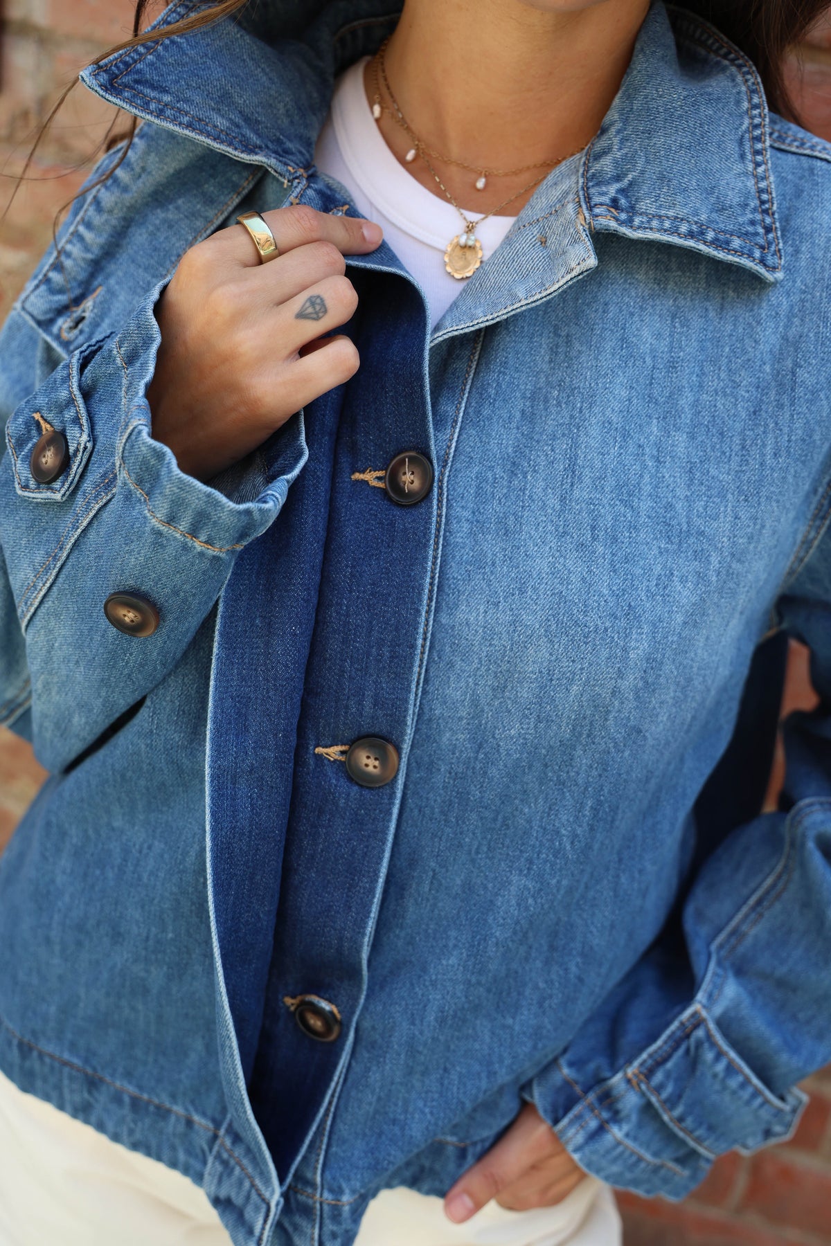 A close-up of a person wearing a blue denim jacket over a white top, with visible hands, neck, jewelry, and part of a brick background.