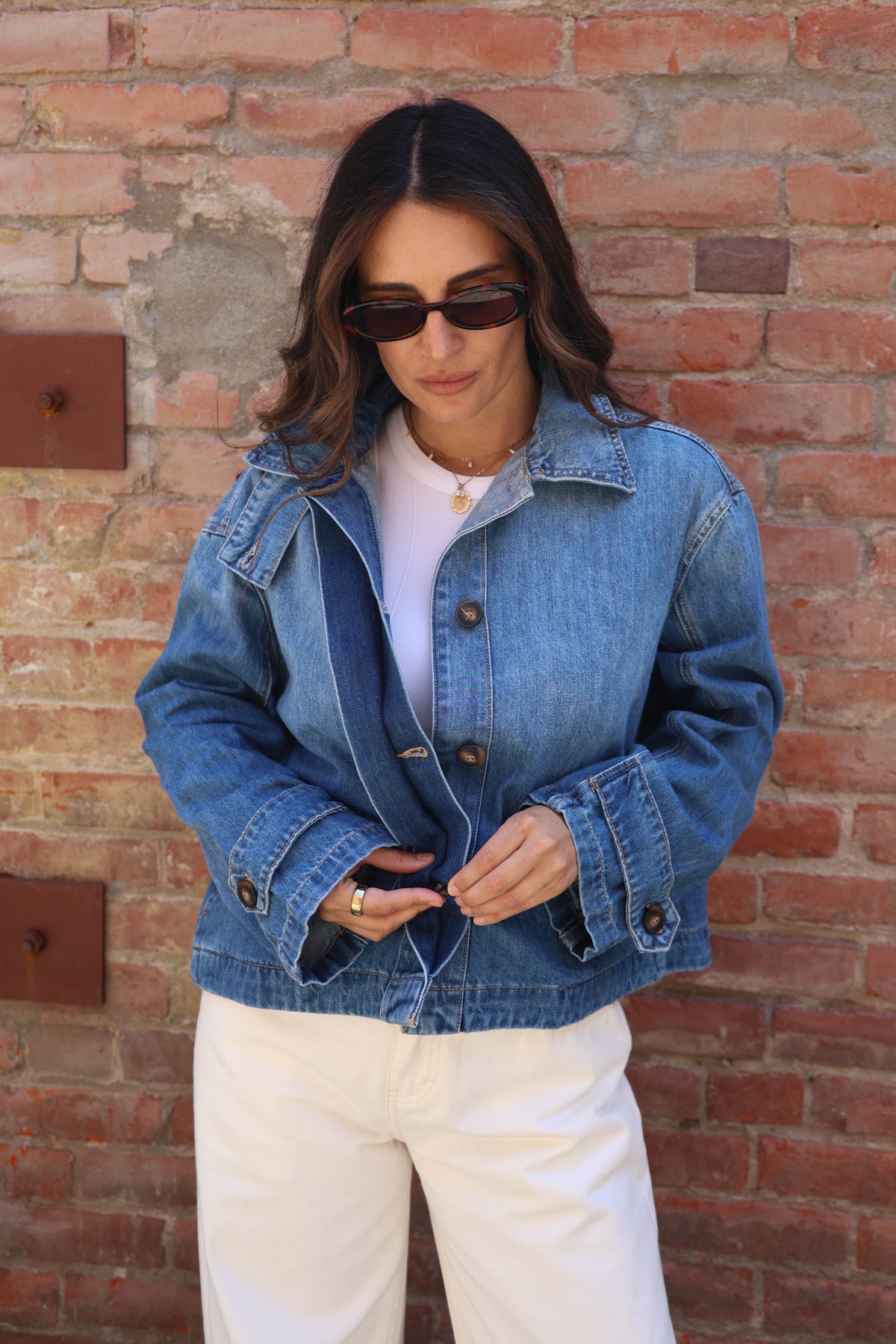 A woman wearing a light-wash blue denim jacket over a white top and white pants, standing in front of a red brick wall.