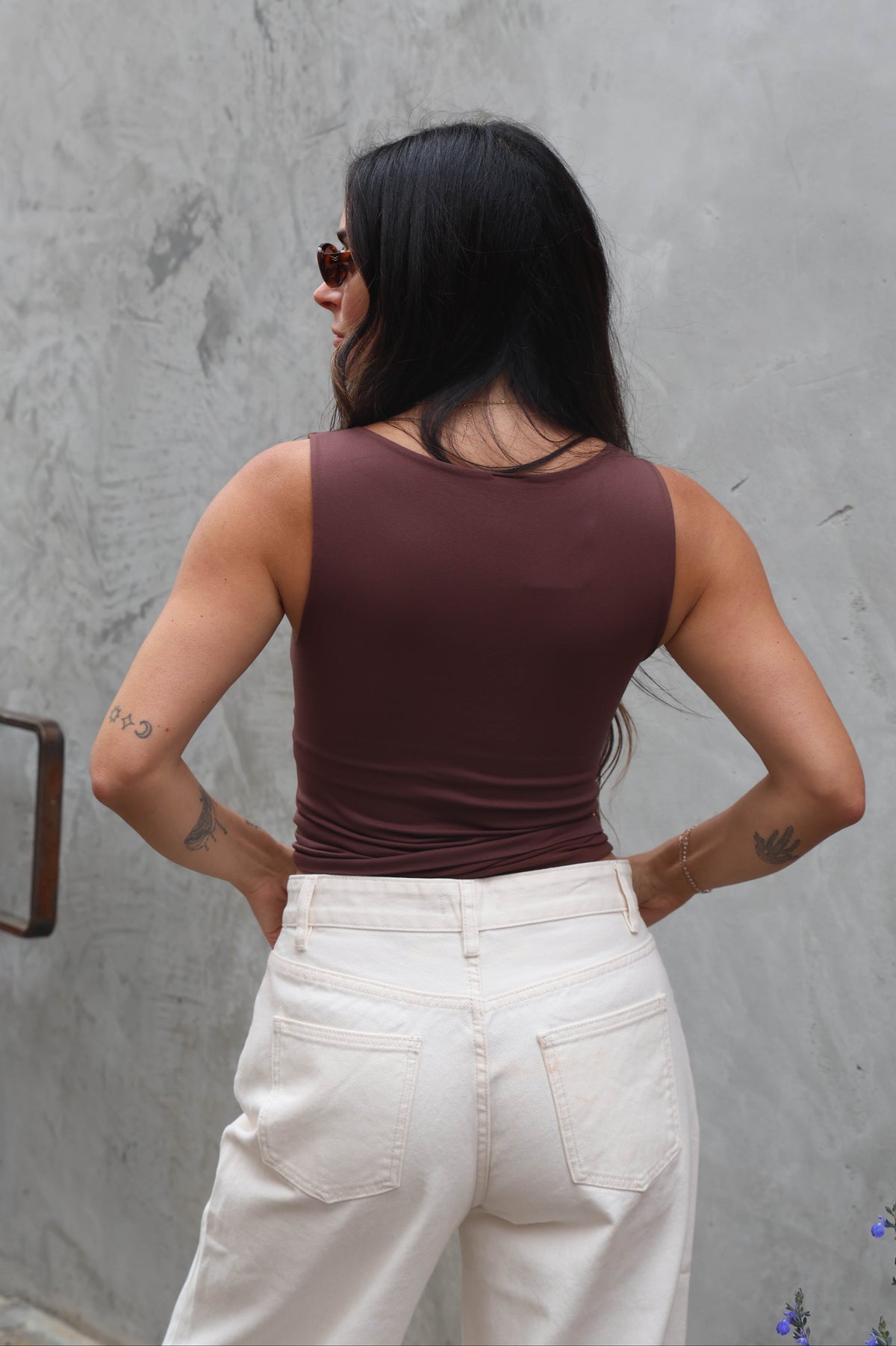 Woman wearing a brown tank top and white pants standing against a gray wall.