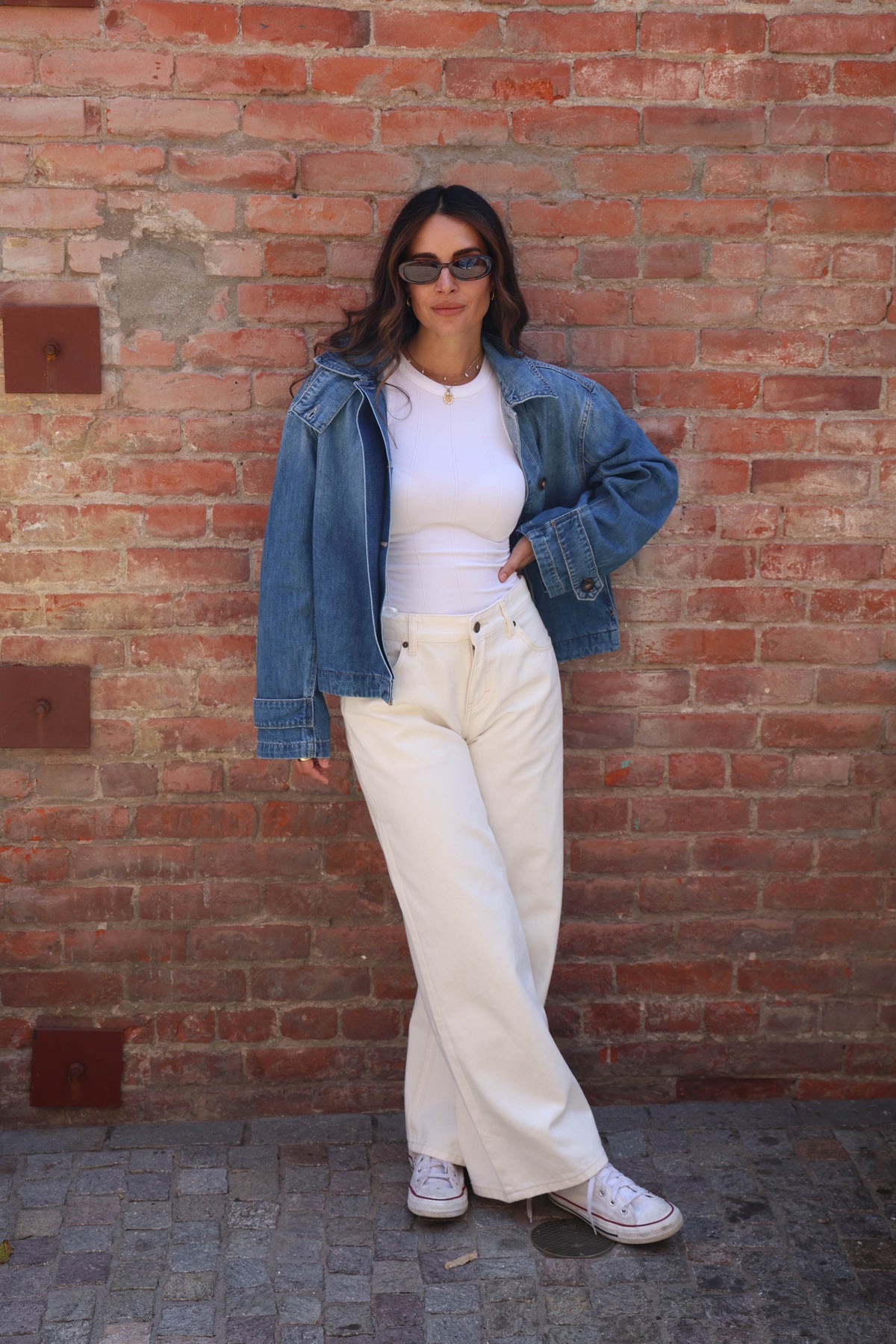 A woman posing in front of a red brick wall wearing a blue denim jacket, white ribbed top, off-white wide-leg jeans, and white sneakers.