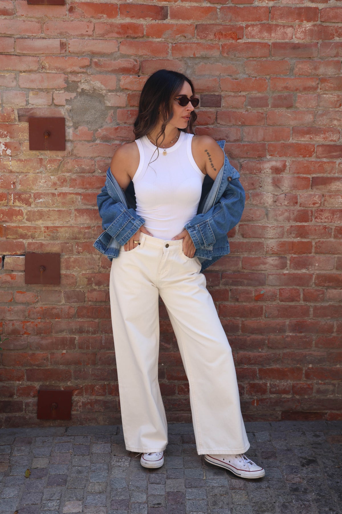 A woman stands against a red brick wall wearing a white ribbed tank top, off-the-shoulder blue denim jacket, cream wide-leg trousers, and white sneakers.
