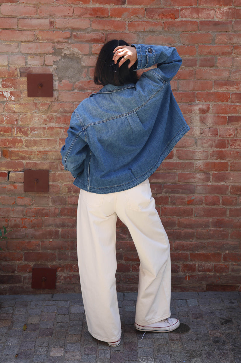 A person standing with their back to the camera in front of a red brick wall, wearing a blue denim jacket, cream wide-leg trousers, and white sneakers.