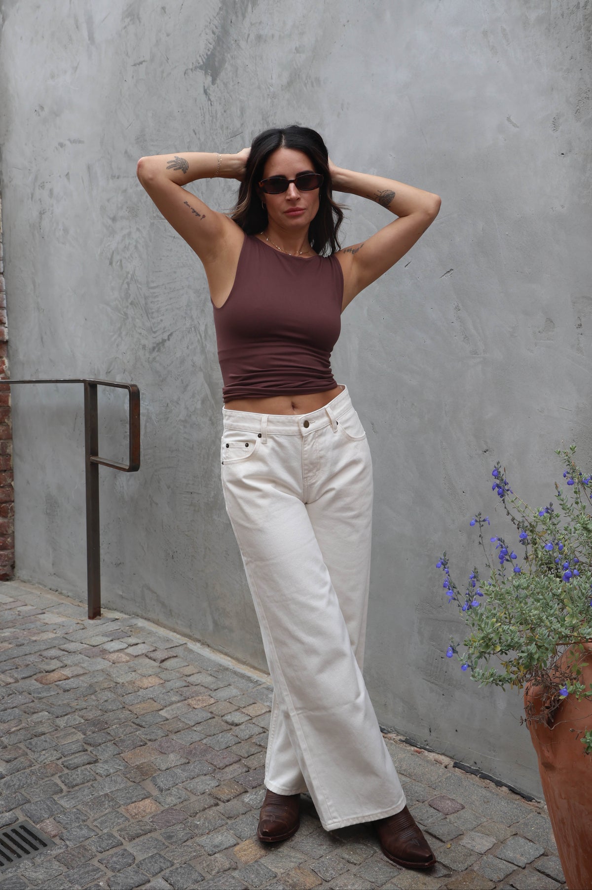 Woman in a brown top and white pants standing against a gray wall with a plant in the foreground.
