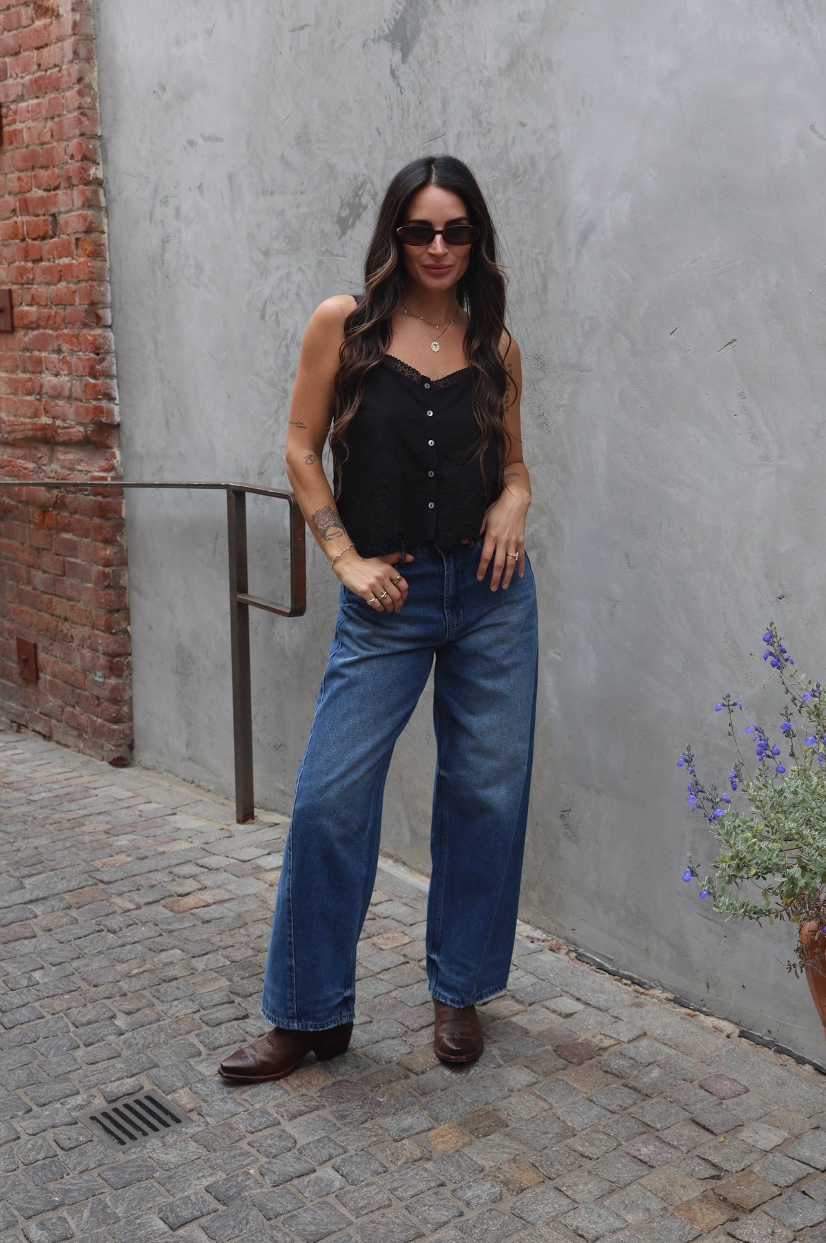 Woman in black top and blue jeans standing against a gray wall with a brick wall in the background.
