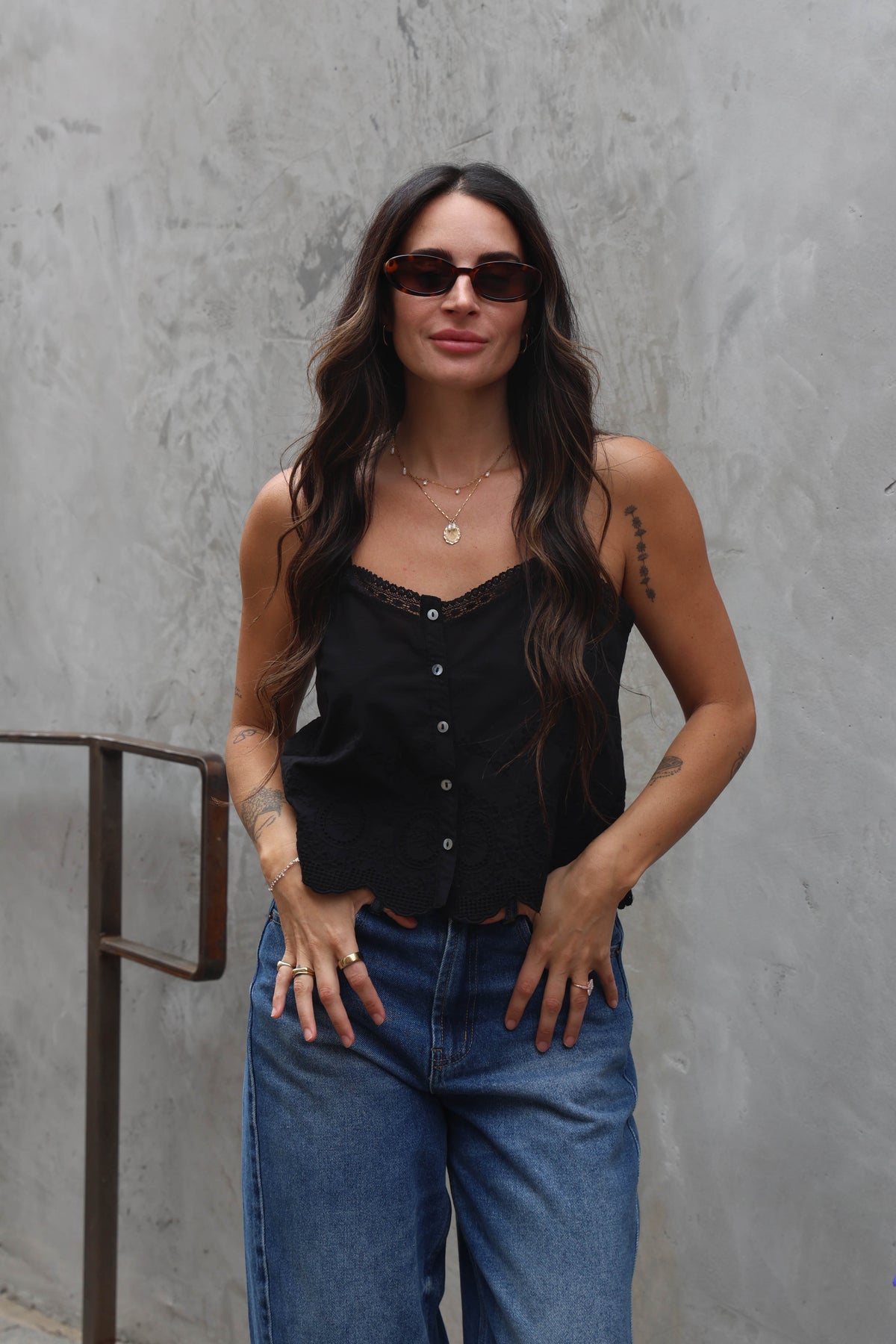 Woman wearing a black tank top and blue jeans against a gray wall.
