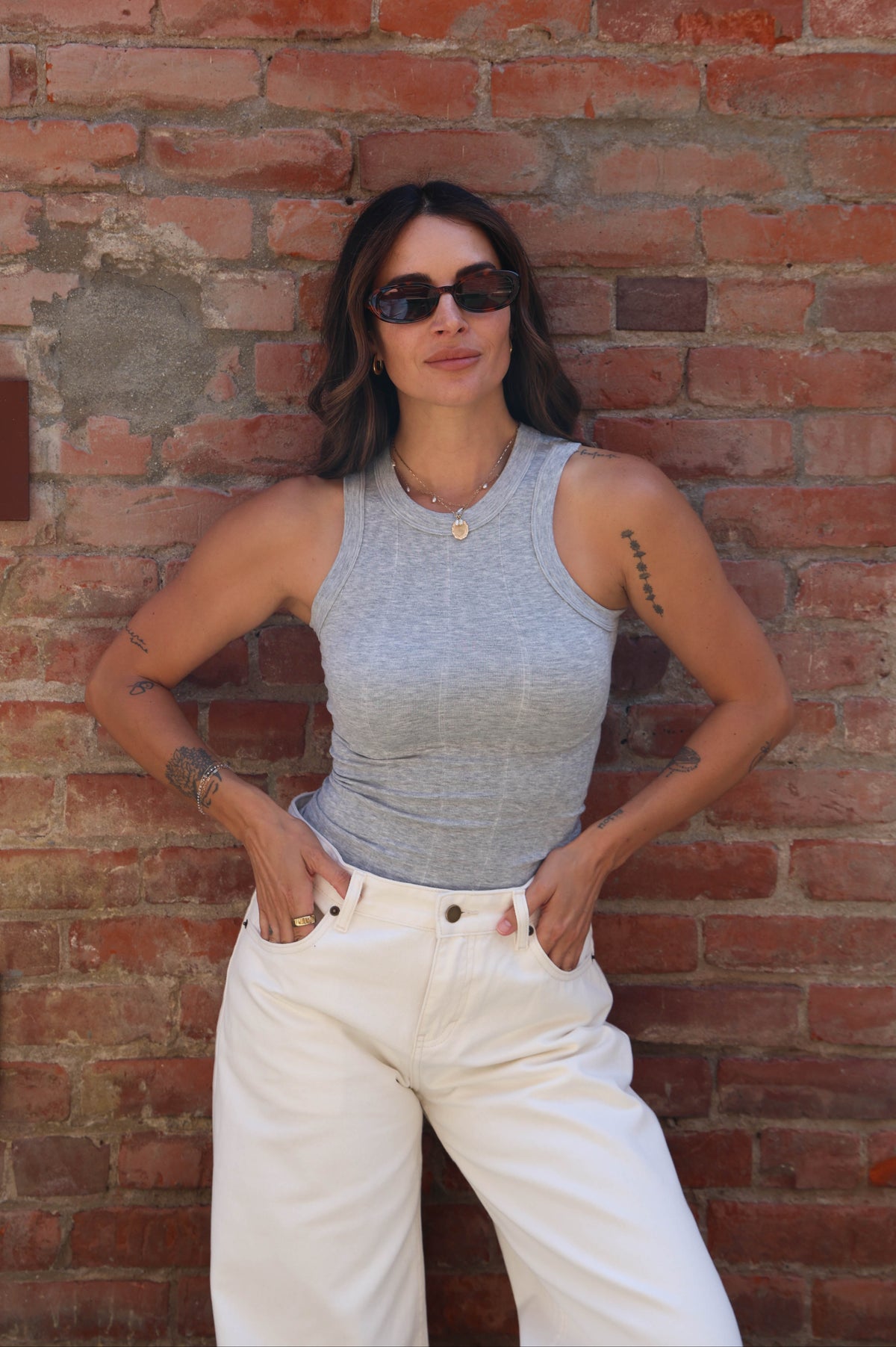 A woman posing against a red brick wall, wearing a fitted heather gray sleeveless tank top and white wide-leg jeans.